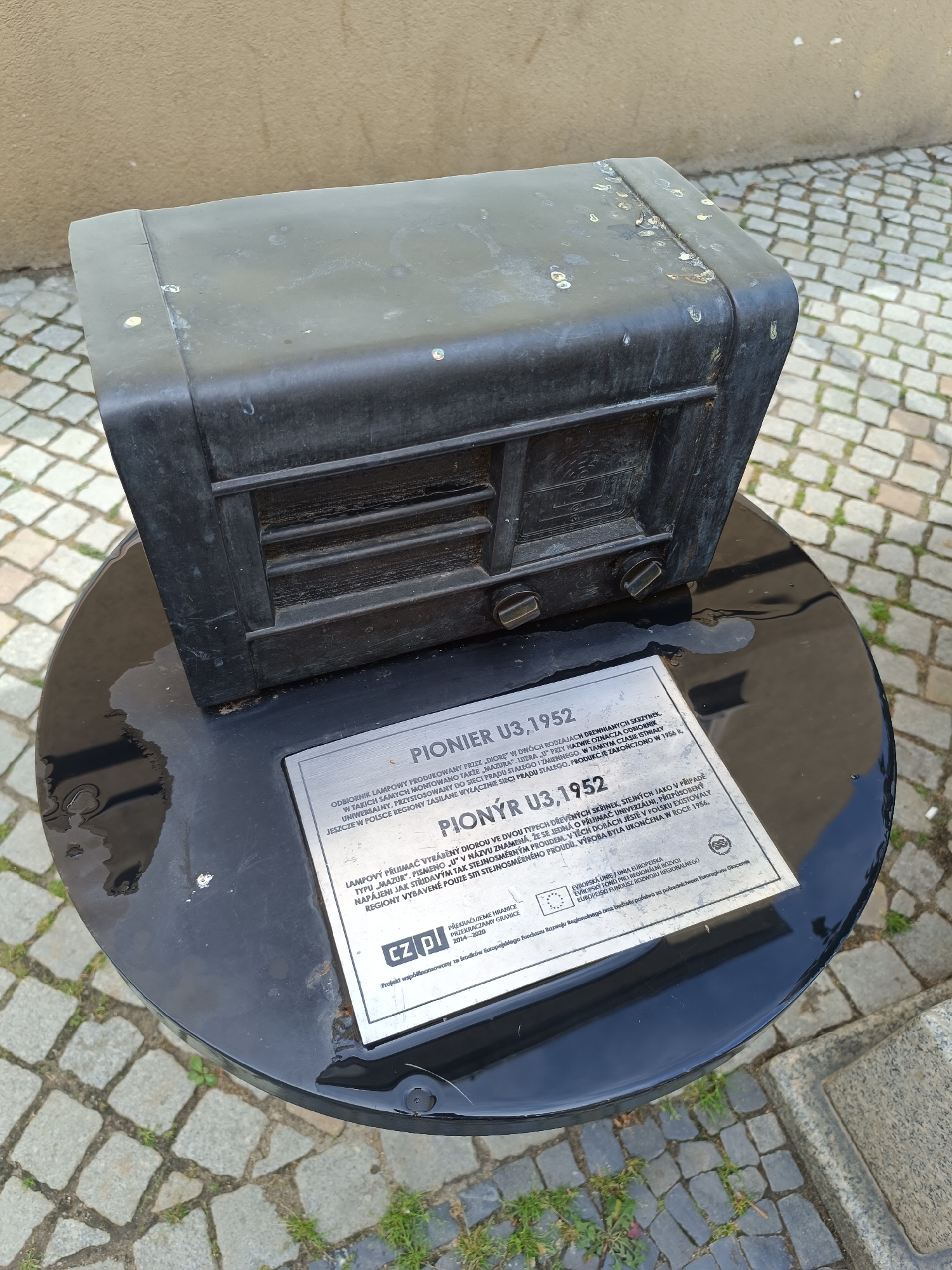 The image depicts a black, weathered metal sculpture of an old radio, specifically a "PIONYR U3,1952," displayed outdoors on a round black pedestal. The pedestal is placed on a cobblestone ground with some grass growing between the stones. A plaque is affixed to the front of the pedestal, providing information about the radio sculpture. The background shows a textured wall and more cobblestone. The sculpture appears to be located in the city of Dzierżoniów, Poland, as indicated by the text on the plaque which mentions "Dzierżoniów" and a project co-financed by the European Regional Development Fund. There are no people visible in the image.