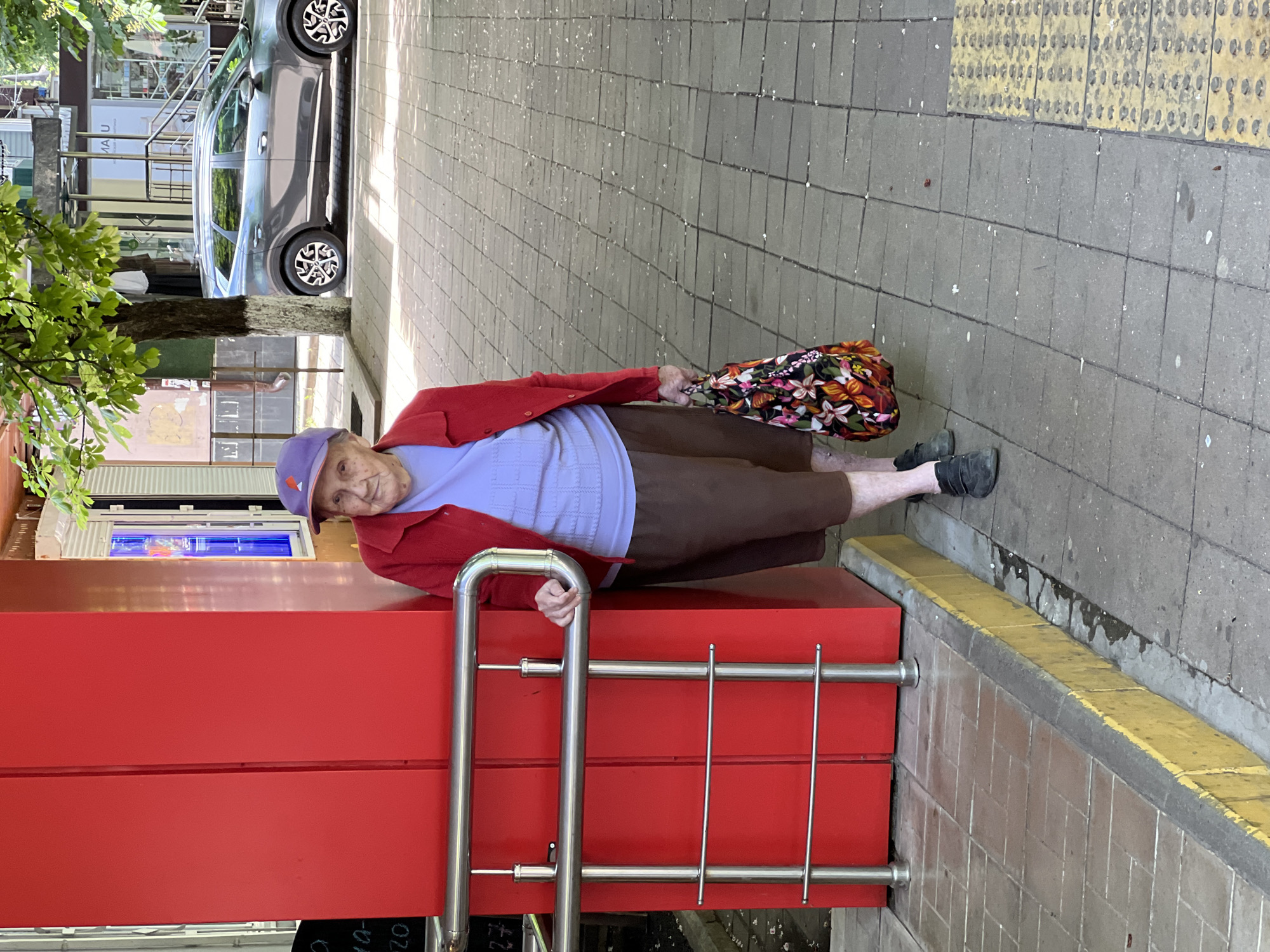An elderly woman in a red cardigan, light purple sweater, brown pants, and a purple cap stands on a paved sidewalk, leaning against a red structure with silver railings. She holds a floral bag in her left hand. The location is Krasnodar, Russia. A car is parked behind the tree on the left side of the frame, and tactile paving is visible on the right.