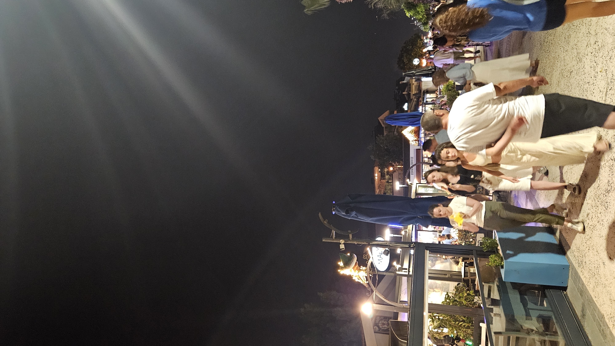 A nighttime scene shows a busy pedestrian street lined with illuminated businesses. Several people are walking along the path. On the left, a restaurant or cafe has tables and chairs visible through a glass facade, with an illuminated sign reading "MALIK." Several blue umbrellas are prominent in the scene, some folded and some open. In the background, buildings are lit up, suggesting a commercial or entertainment district. The sky above is dark with streaks of light, possibly from streetlights or distant sources. The overall impression is of a lively evening atmosphere in an urban setting, likely a coastal town given the presence of umbrellas and the casual attire of the pedestrians. This appears to be a scene from the Paspatur Çarşısı (Old Town) in Fethiye, Turkey, a popular area known for its shops, restaurants, and nightlife.