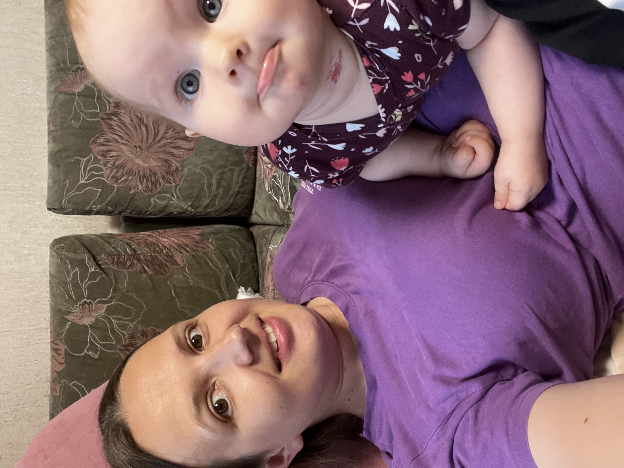 In this image, a woman and a baby are lying down, likely on a couch, given the floral patterned fabric visible in the background. The woman is wearing a purple t-shirt and appears to be looking directly at the camera, with a slight smile. Her eyes are brown, and her hair is dark.
The baby, positioned above the woman, is looking upwards with wide, blue eyes. The baby is wearing a dark-colored onesie with a floral print. There's a noticeable reddish mark on the baby's chin and neck area. The lighting suggests it is daytime, and there are no strong indicators of the weather or specific time of day beyond that. The overall emotion conveyed is one of affection and playfulness between the woman and the baby. The setting seems to be indoors, possibly a living room, given the furniture and the casual nature of the pose. There is no visible text in the image.