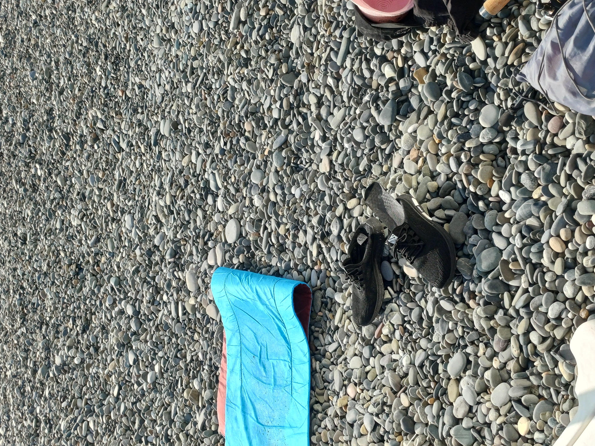 A close-up, overhead shot shows a pile of smooth, gray and beige pebbles covering a beach. A bright blue sleeping bag, partially unrolled and revealing a reddish-brown interior, lies on the pebbles in the upper left quadrant. To the right of the sleeping bag, a pair of black athletic shoes are placed next to each other. The shoes appear to be mesh with black laces. A black sock or fabric is visible underneath the right shoe. To the far right, partially visible, is a pink object, possibly a yoga mat, and some dark fabric. The scene is set on a beach, likely in Sochi, Russia, given the characteristic pebble-strewn shorelines common in the area. The lighting suggests it is daytime, with shadows cast by the objects indicating a slightly angled sun. No people are visible in the frame, but the presence of personal belongings suggests recent activity.