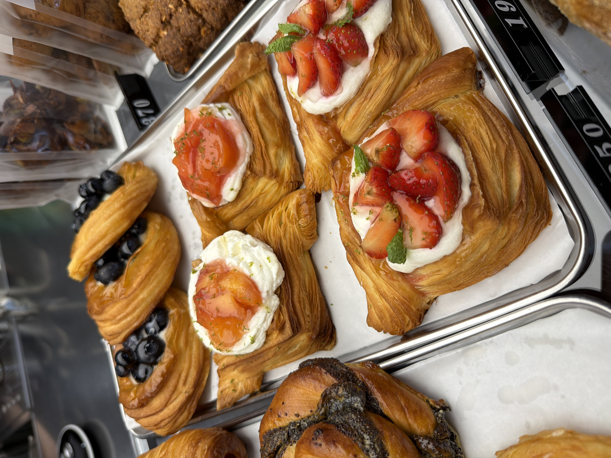 The image shows a close-up view of a display of pastries in a bakery. There are several types of pastries visible, including Danish pastries topped with blueberries and cream, and pastries with strawberries and cream. A pastry with a poppy seed swirl is also present. The pastries are arranged on a metal tray, which is sitting on a counter. In the background, some packaged goods and a price tag with the number "370" are visible. Another price tag with the number "1950" is seen on the lower right side of the image. The lighting is bright, highlighting the textures and colors of the pastries. The setting appears to be a retail environment, likely a bakery or cafe. There is no information in the image to indicate the specific location in Izhevsk, Russia.