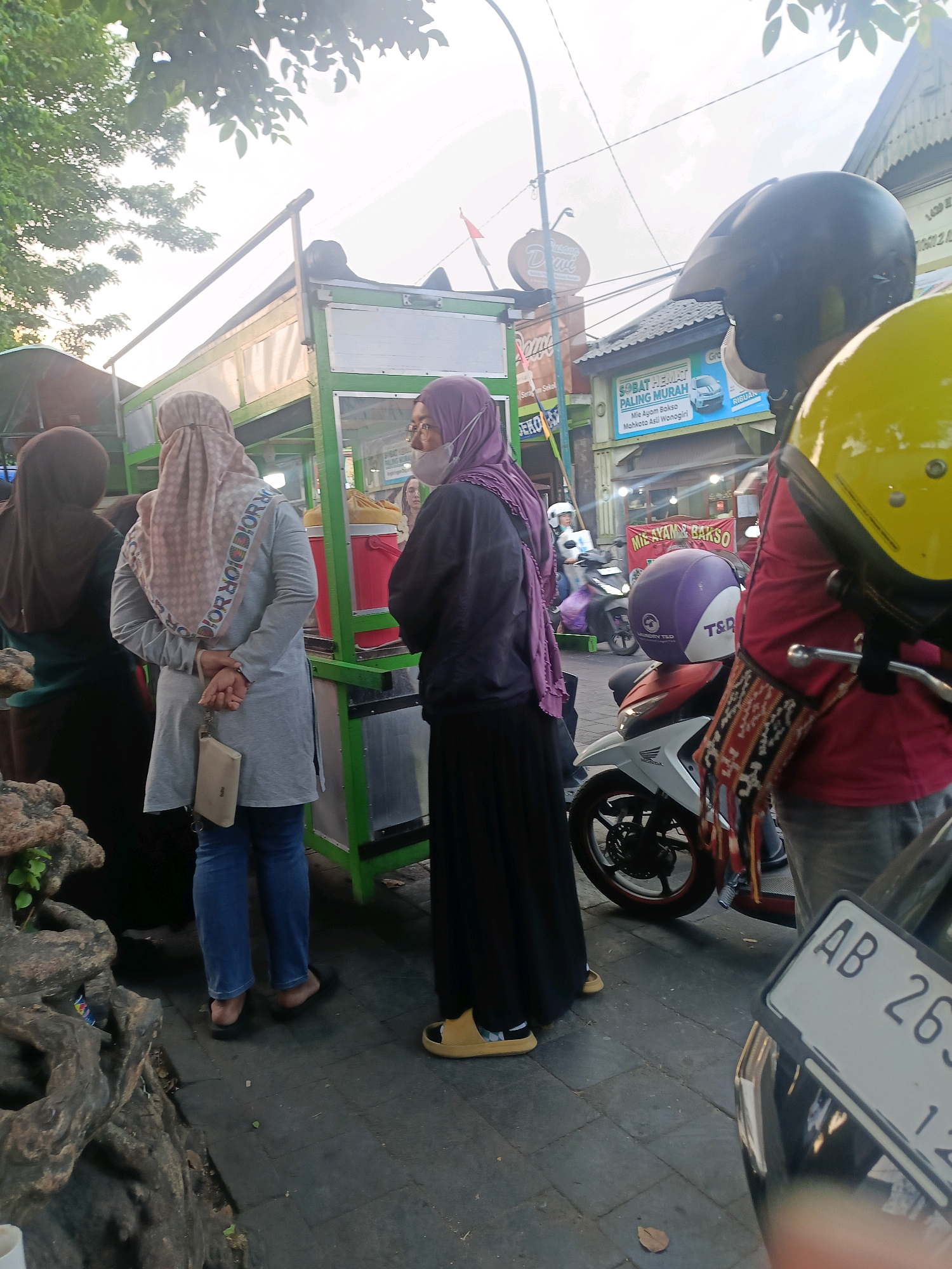 The image depicts a street scene in Yogyakarta, Indonesia, likely near a food stall. Three individuals are partially visible, all wearing headscarves. The person in the foreground, facing away, wears a light gray tunic and blue jeans, with a small beige clutch bag hanging from their left hand. To their left, another person is mostly obscured, wearing a dark brown headscarf and dark clothing. To the right, a third person, seen from the side, wears a dark purple headscarf and a black long skirt, with a visible face mask.
The background features a green food stall with a transparent display case, suggesting food items are being sold. Signs in Indonesian are visible, including one that reads "MIE AYAM BAKSO". Various motorcycles are present, including one with a yellow helmet prominently displayed and another with a purple and white helmet. The ground is paved with dark tiles. The overall lighting suggests it is either early morning or late afternoon.