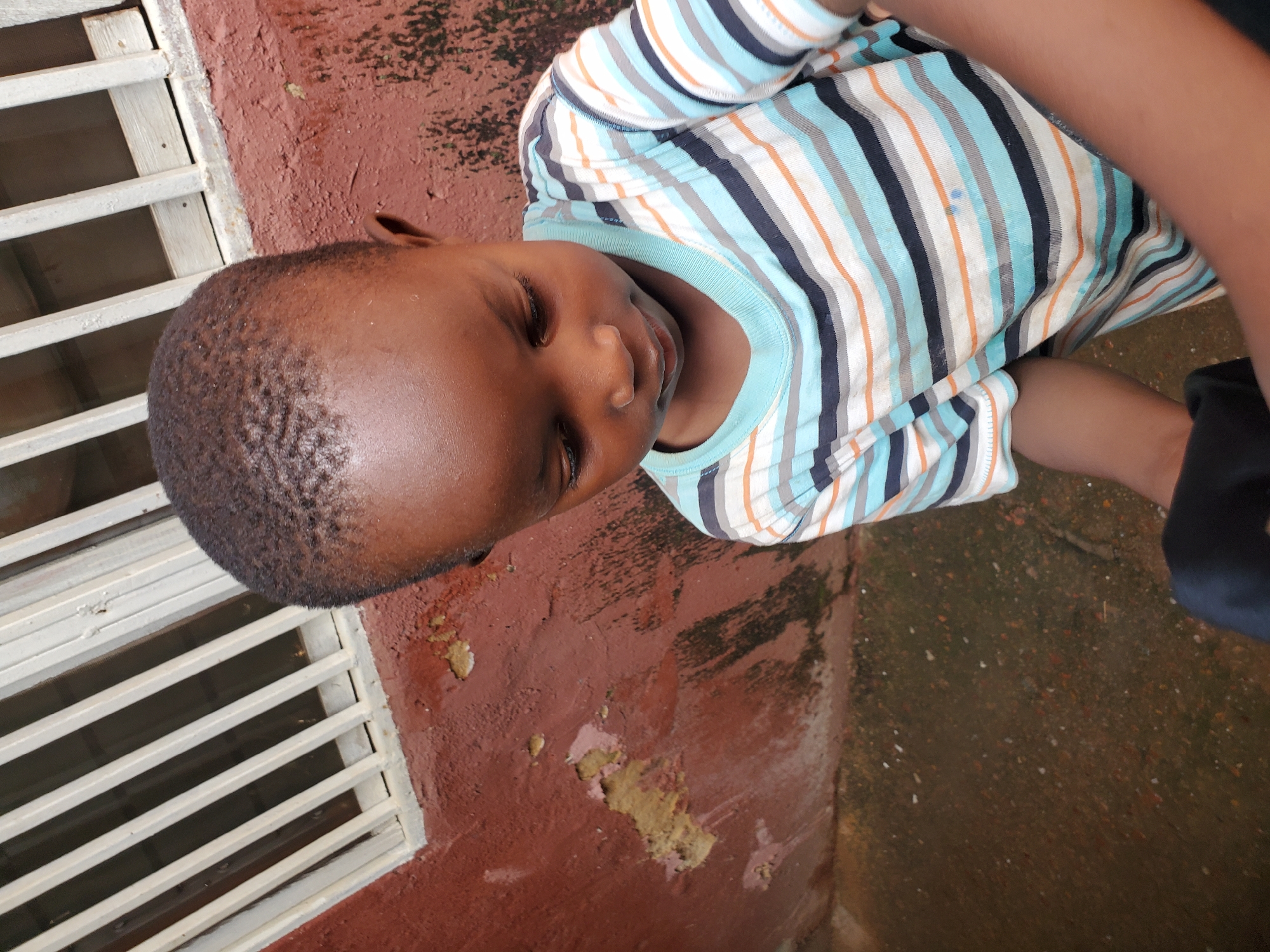 The image is an outdoor, close-up shot of a young child wearing a horizontally striped t-shirt with blue, orange, and dark blue stripes. The child is looking down with closed eyes, appearing relaxed or asleep. The child's head and upper body are in focus, with their arm visible on the right side of the frame.
The background consists of a reddish-brown textured surface, possibly concrete or a wall, with some scattered debris and lighter colored patches that might be paint or residue. To the left of the child, there's a portion of a white, slatted structure, possibly a window grill or vent, suggesting an urban or built environment. The ground in the lower right of the image is darker and appears wet or damp, with small specks of lighter material scattered across it.
The lighting suggests it is daytime, with natural light illuminating the scene. No specific time of day or weather conditions can be definitively determined, although the damp ground might indicate recent rain. There are no visible text elements in the image. The overall scene depicts a moment of quiet repose for the child in an outdoor setting, possibly near a building. The cropped nature of the image and the child's posture do not provide explicit cues about the activity or event.