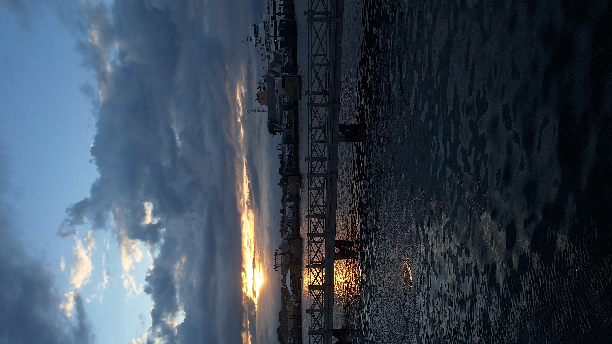 The image captures a view of Nantucket Harbor at sunset, with the sky filled with dramatic clouds that are beginning to clear, allowing the sun to peek through, casting a warm glow on the water and the distant shore. In the foreground, a pedestrian bridge with a crisscross railing spans the water. The water itself is dark and reflects the fading light, with small ripples distorting the reflections.
The mid-ground shows the Nantucket waterfront. Several buildings are visible, likely part of the town's commercial or residential areas. A large ferry, the Iyanough, is docked or moored, its white hull and superstructure prominent against the darkening sky. Other smaller vessels and docks are also present. The sun, low on the horizon, is partially obscured by clouds, creating a dramatic lighting effect with streaks of orange and yellow light breaking through. The clouds themselves are a mix of dark grays and lighter tones, adding depth and texture to the sky. The overall atmosphere is serene and evocative of the end of a day in this historic island town.