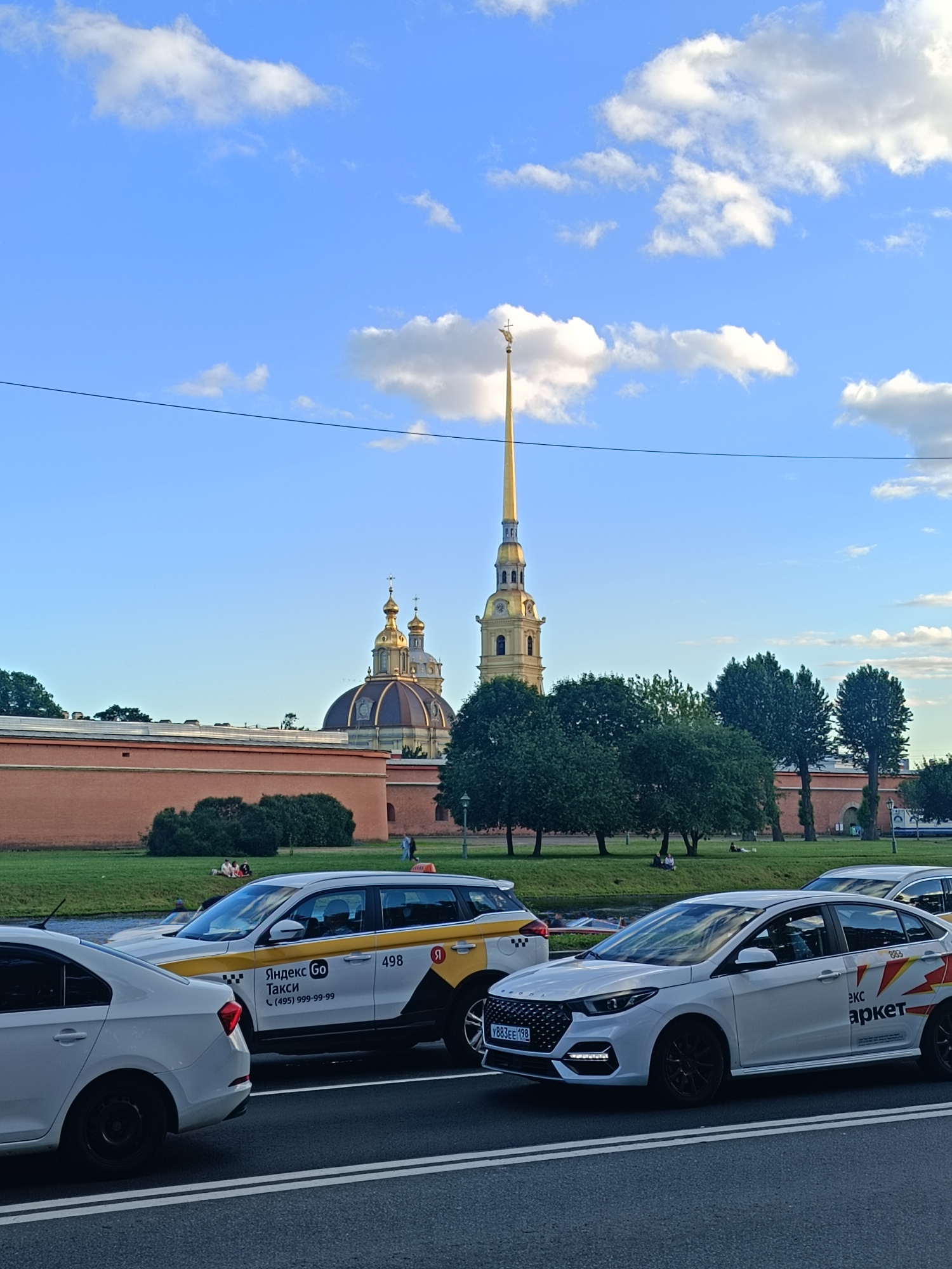 In the city of Saint Petersburg, Russia, on a cloudy day, the Peter and Paul Fortress stands prominently. A golden spire tops a tall bell tower, flanked by domed cathedrals with golden roofs. The fortress is surrounded by a red brick wall, and lush green trees line the perimeter.
In the foreground, several white cars are parked on a street, some of which are marked with the Yandex Go taxi service logo. The cars are a mix of sedans and SUVs, and they are all facing in the same direction. The street is paved with asphalt, and there are white lines painted on it to indicate lanes.
In the background, a body of water separates the street from the fortress. A few people are seen sitting on the grass near the fortress wall, enjoying the scenery. The sky is blue with scattered white clouds, and the overall atmosphere is peaceful and serene.