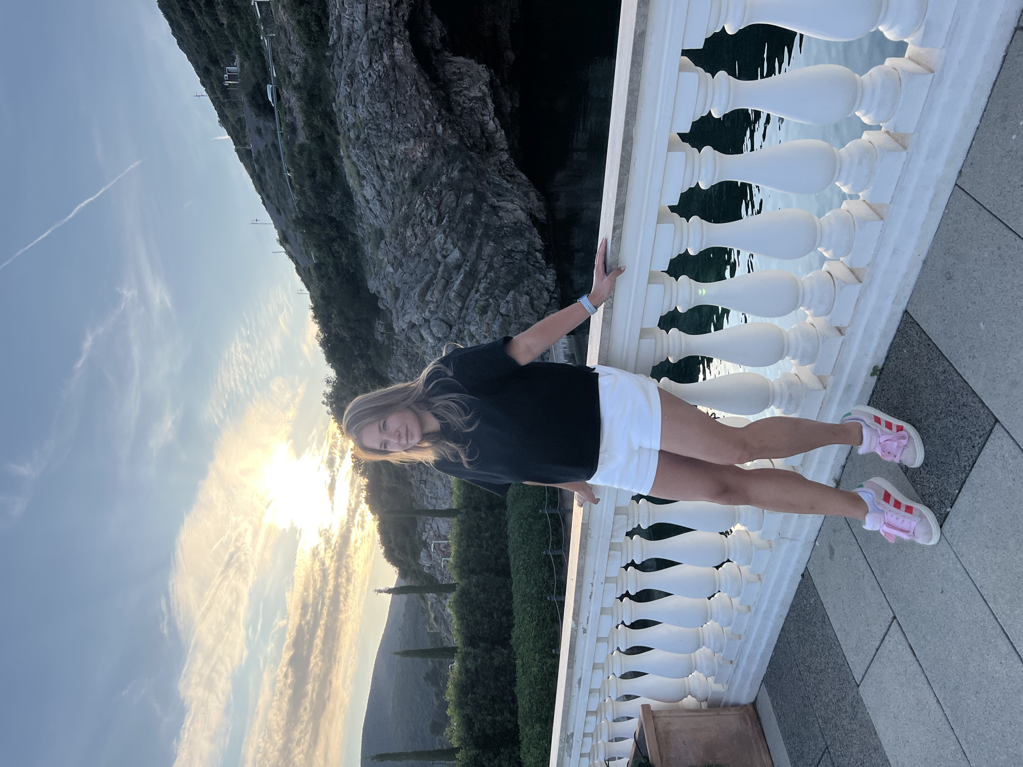The image, correctly oriented, captures a young woman posing on a scenic outdoor balcony or viewpoint overlooking a coastal landscape during the late afternoon or early evening, likely at sunset. The setting is consistent with Radovići, Montenegro.
The primary subject is a young woman with long blonde hair, dressed in a black t-shirt, white shorts, and pink and white sneakers. She leans casually against a white stone balustrade, looking towards the camera with a subtle smile, appearing relaxed and content. A blue wristband is visible on her left wrist.
The background reveals a dramatic and beautiful natural environment. To the left, dark, rugged rocky cliffs descend steeply towards a body of water, which appears to be the sea or a large bay. The water visible below the balustrade is calm and reflects the surrounding elements. To the right, green, tree-covered hills and mountains extend into the distance, with several tall, slender cypress-like trees dotting the landscape, enhancing the Mediterranean feel.
The time of day is indicated by the brilliant golden sunlight, which is low in the sky, creating a warm glow. The sky transitions from a clear blue to softer, cloud-streaked areas illuminated by the setting sun. The woman stands on a light grey paved surface. There is no visible text in the image.