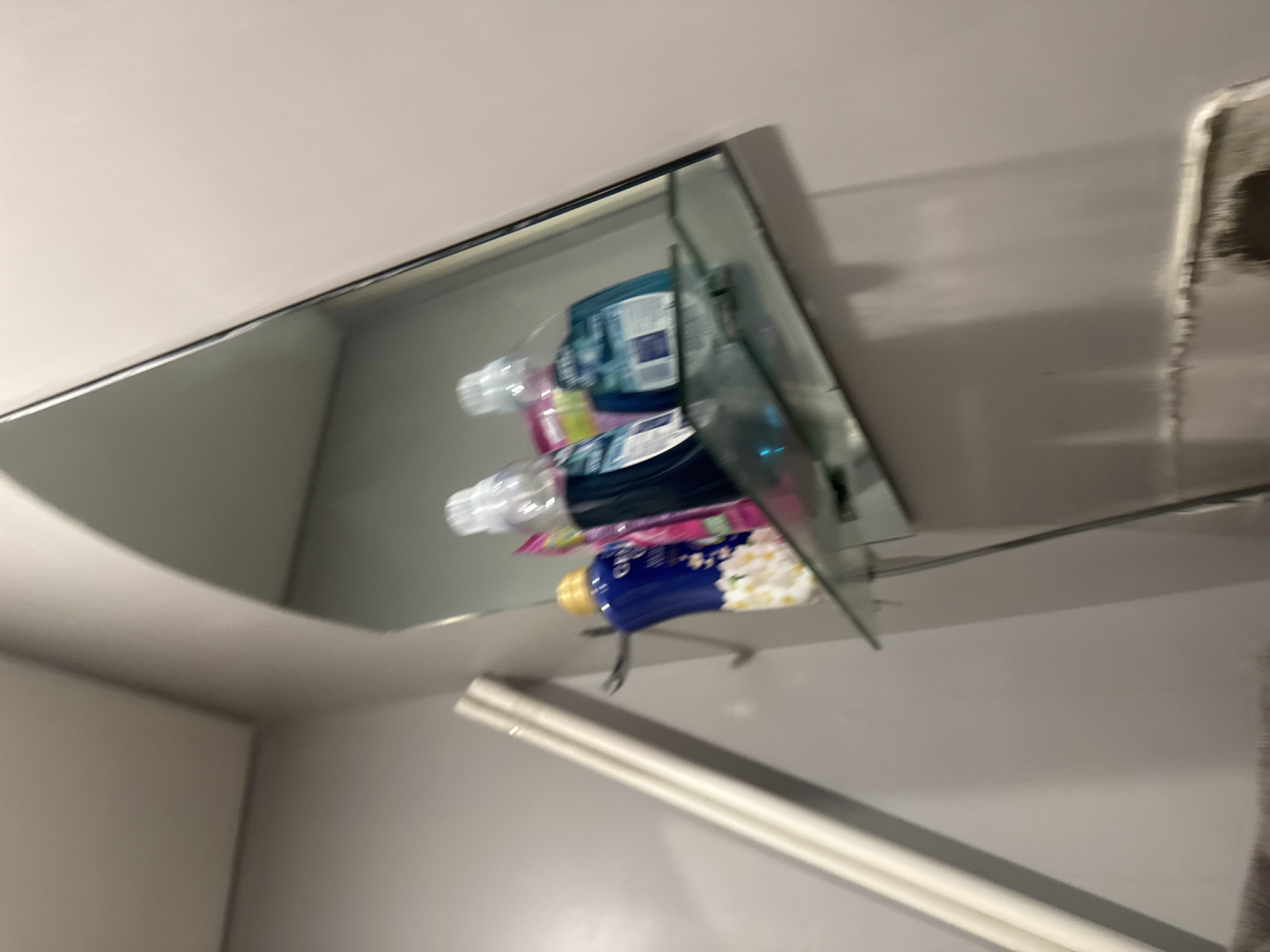 The image shows a close-up, slightly blurry view of a shelf with various bottles and containers. The shelf appears to be made of glass and is mounted on a wall. The items on the shelf include what look like toiletries, such as bottles of lotion or soap, and possibly mouthwash. Some of the bottles have colorful labels, and one appears to be a blue bottle with a yellow cap. The background is a plain wall, and there is a large mirror reflecting the items on the shelf, creating a sense of depth. The lighting is somewhat dim, and the overall impression is of a bathroom or vanity area. There is a long, white object running along the bottom of the frame, possibly a towel rack or a cabinet. The image is taken from a slightly upward angle.