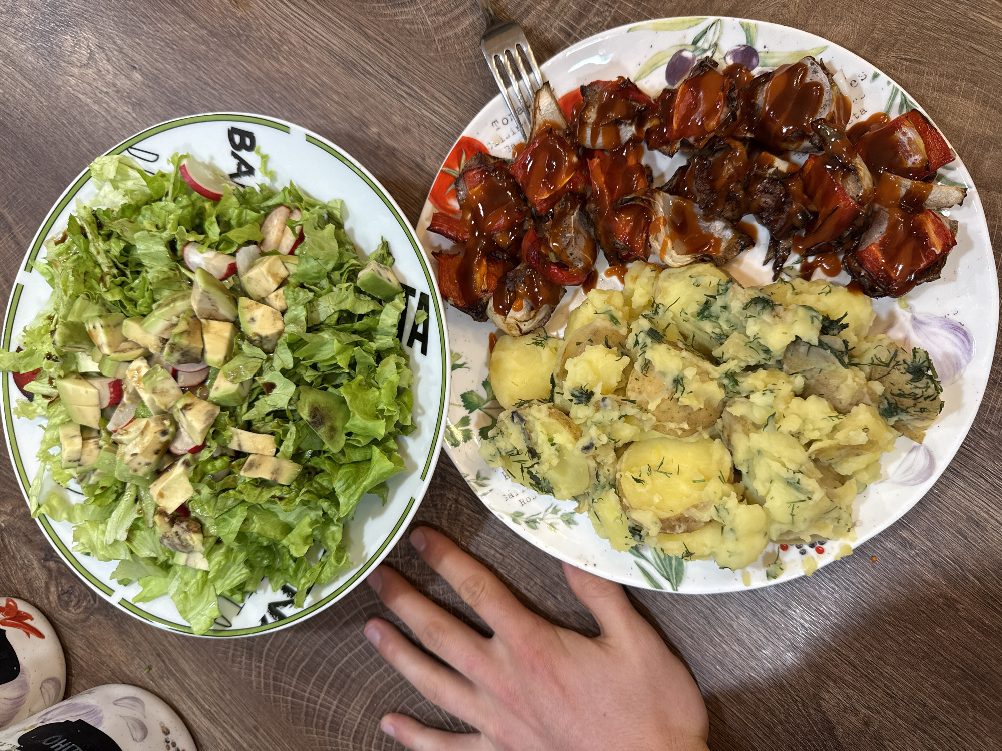An overhead shot captures a home-cooked meal served on a wooden table, likely in a domestic setting in Sofia, Bulgaria. The meal consists of two main dishes presented on separate plates.
The plate on the left holds a fresh green salad made primarily of torn lettuce leaves, cubed avocado, and slices of red radish. A dark, possibly balsamic, dressing is drizzled over the salad. This plate is white with a thin green border and features black text, partially visible as "BA" and "D".
The larger plate on the right presents a main course with two components. The upper section contains grilled items, appearing to be pieces of meat (possibly pork or chicken) and red bell peppers, heavily coated in a rich, dark red barbecue sauce. Below this, a generous portion of boiled or lightly mashed potatoes, visibly seasoned with chopped fresh green herbs like dill or parsley, occupies the lower part of the plate. A silver fork rests on the upper left side of this plate. The plate itself is white and decorated with illustrations of tomatoes, olive branches, garlic, and text fragments such as "Tomato," "Cuisine," "TomaTA," "es," "ta," "Galli," and "Ros."
A light-skinned hand, presumably a male's, is visible on the wooden table surface, reaching towards the right plate, indicating an imminent or ongoing meal. The table has a warm, medium-brown wooden finish with distinct grain patterns. A portion of patterned, possibly ceramic or fabric, footwear is visible on the bottom left corner of the image. The scene conveys a casual, ready-to-eat meal.