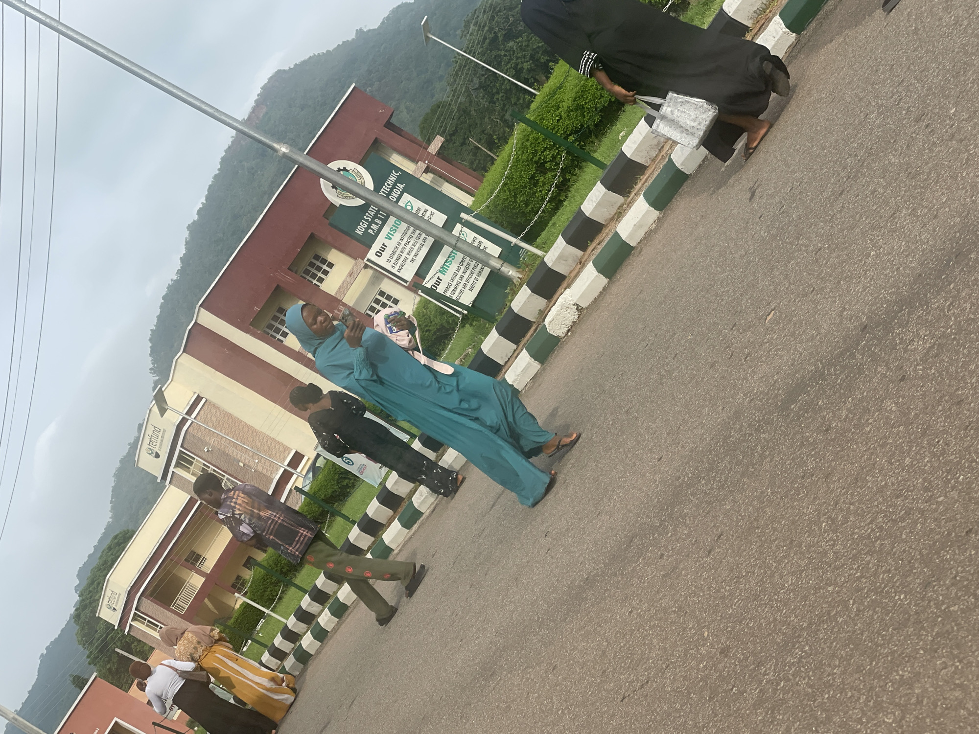 A group of people walk along a paved road in front of a building with the inscription "KOGI STATE POLYTECHNIC, lokoJA." The building has a red and beige facade with multiple windows. A sign with "Our Vision" and "Our Mission" is visible on a green pole. People are dressed in traditional Nigerian attire. One person in a teal hijab is looking at their phone. Another person in a black abaya is carrying a silver tote bag. A person in green pants and a patterned shirt walks away from the camera. Two people in the background are wearing yellow and brown attire. The sky is overcast with a view of a mountain in the background.