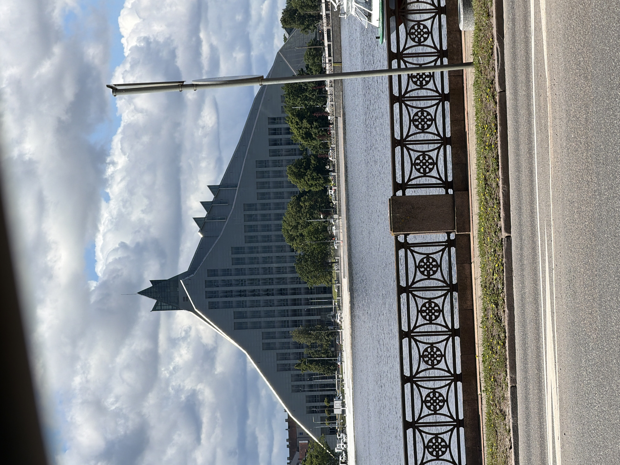 The image shows the Latvian National Library in Riga, Latvia. The library is a modern, triangular-shaped building with a distinctive pointed roof. It is situated on the bank of the Daugava River, with a promenade and a decorative fence in the foreground. Trees line the riverbank behind the fence, and the sky above is filled with dramatic, cloudy formations. A street and a sidewalk are visible in the lower portion of the image, suggesting a public space within the city. A lamppost stands to the right of the building, and a small boat is docked in the river.
