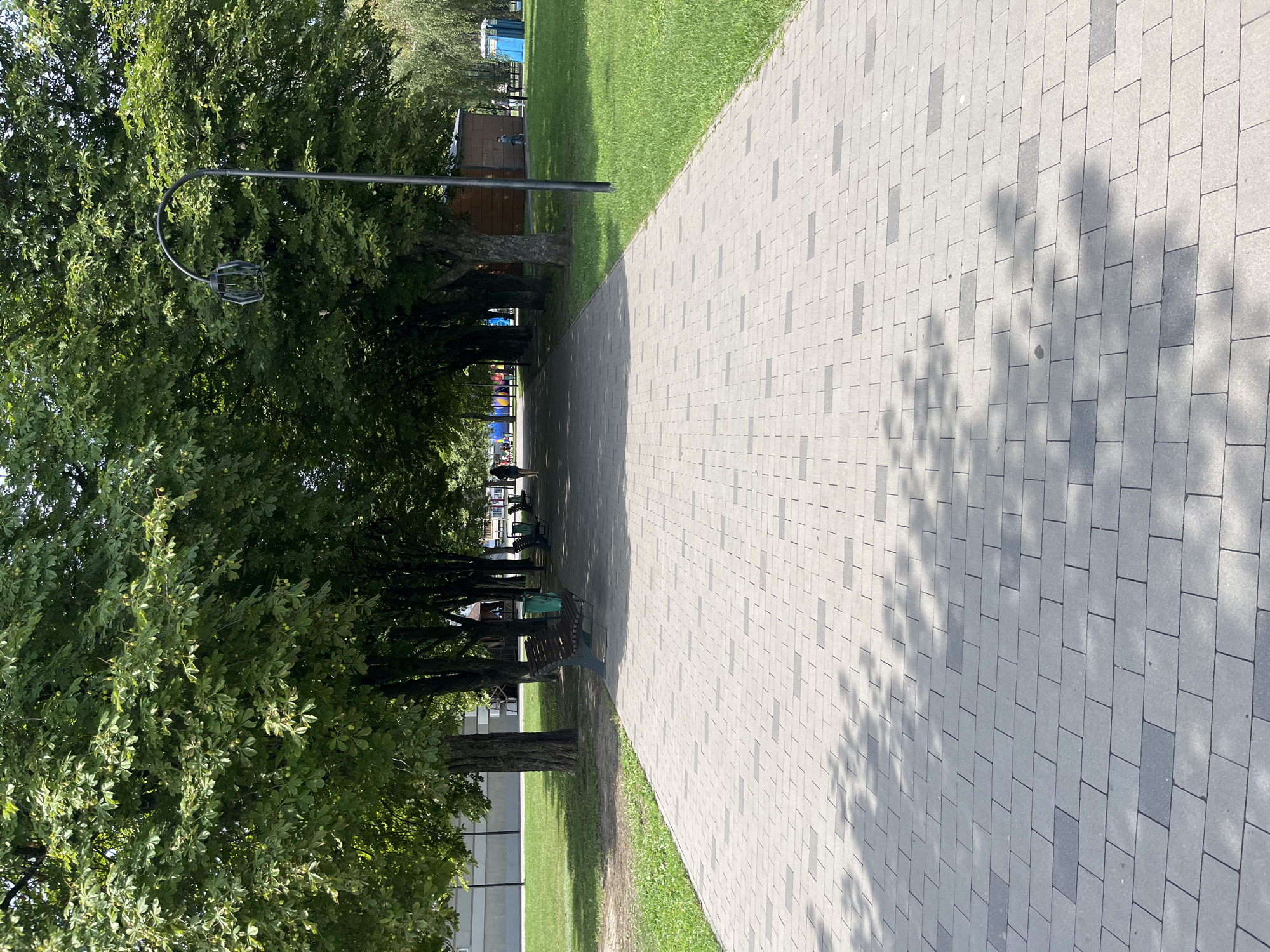 A paved walkway lined with trees leads into the distance in a city park. The walkway is made of light gray and dark gray rectangular pavers arranged in a repeating pattern. On the left side of the walkway, a grassy area borders the path, with a row of trees casting dappled shadows on the pavers. Further into the park, there are benches, trash cans, and a lamppost. On the right side of the walkway, more grass and trees are visible, along with a small wooden building and a blue portable toilet. In the distance, a few people can be seen walking and sitting. The overall scene suggests a pleasant day in an urban park. The location is in the city of Kharkiv, Ukraine.