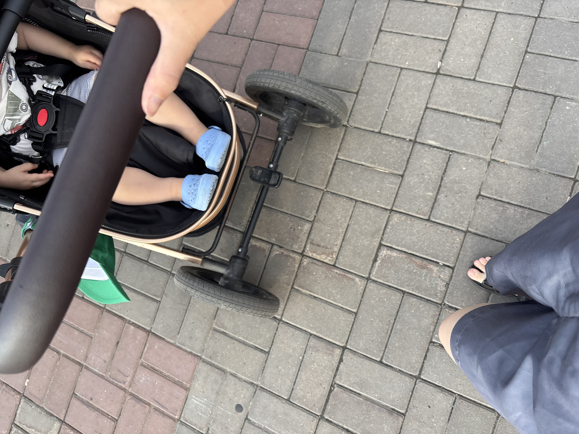 The image shows a top-down view of a stroller with a child sitting in it. A person's hand is holding the stroller's handle. The child is wearing blue Crocs. The stroller is on a paved walkway. In the lower part of the image, the legs and feet of the person holding the stroller are visible, wearing dark shorts and sandals. The environment appears to be outdoors, with interlocking brick paving stones. There are no specific landmarks or indicators to pinpoint the location to Naberezhnye Chelny, Russia, or any other specific city.