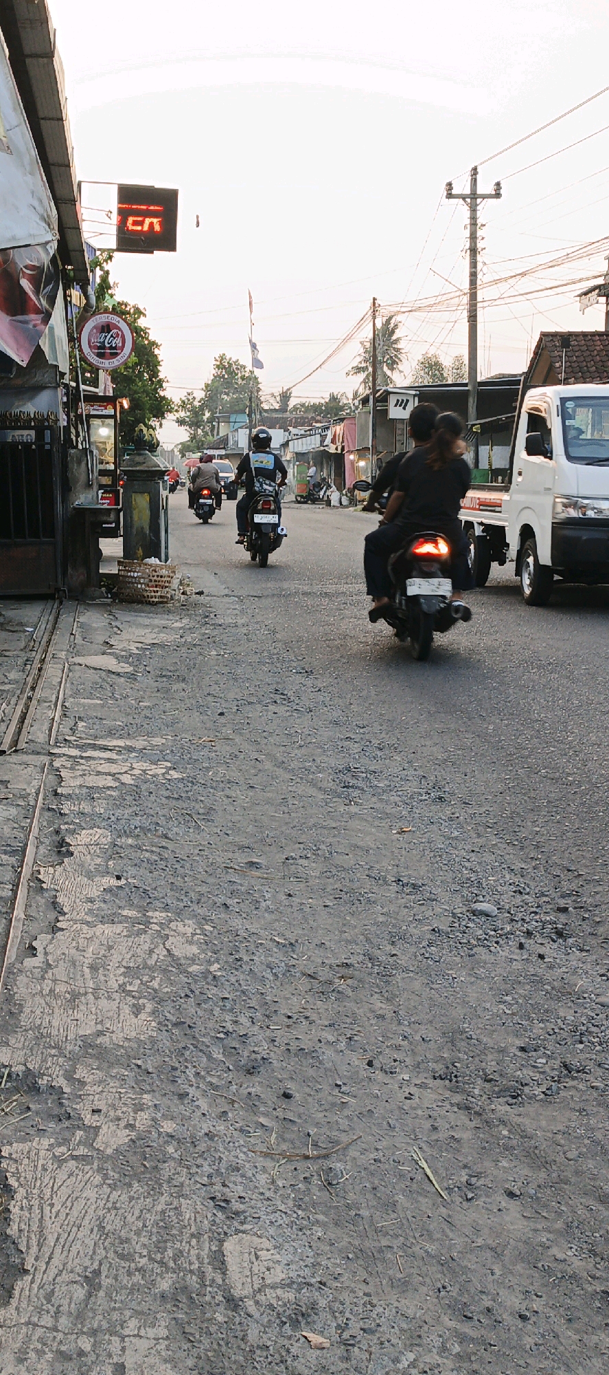 The image depicts a street scene in Sedayu, Indonesia, likely during late afternoon or early evening. The road is paved with a rough, gravelly surface, showing signs of wear and tear. On the left side of the road, a building with a corrugated metal roof and metal gate is visible, with a Coca-Cola sign and a red digital sign reading "TCR" hanging above. Further down the street, several motorcycles are in motion, carrying passengers. One motorcycle in the foreground has two individuals riding it, with the rear passenger leaning forward. Another motorcycle ahead of them also carries two people. On the right side of the street, a white truck is parked or driving slowly. Power lines and utility poles are strung across the sky, characteristic of the area. The background shows residential buildings and some vegetation, including palm trees. The overall atmosphere suggests a typical street in a non-urbanized area of Indonesia, with active local transportation and commercial activity.