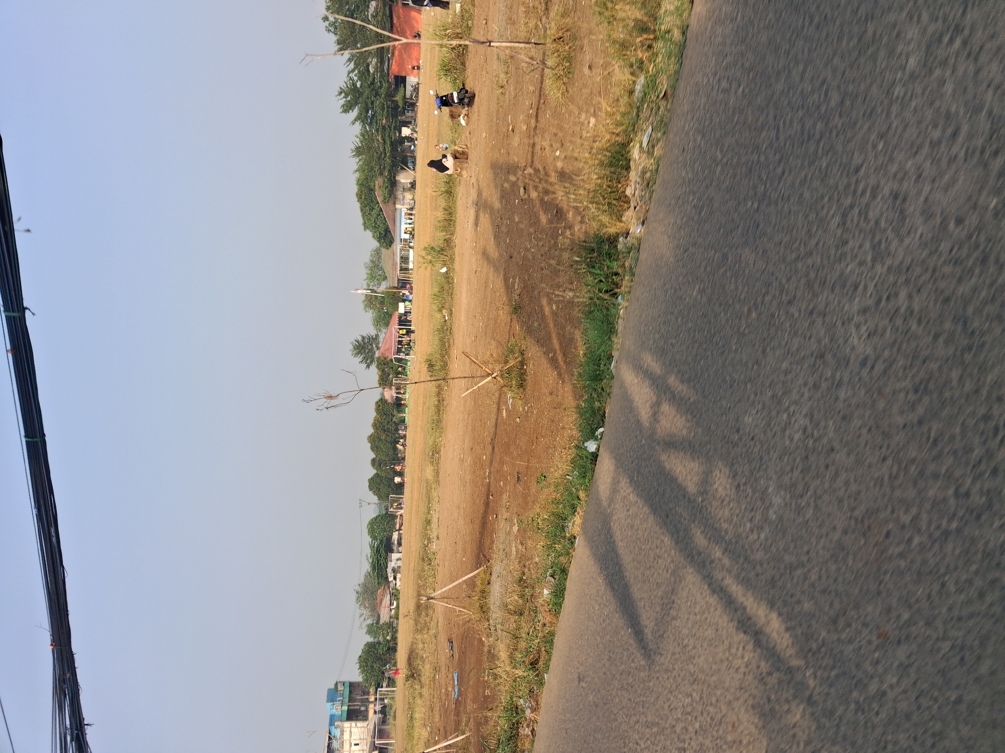 This is a daytime, eye-level, wide-angle shot of an open field area in Bojong Gede, Indonesia, likely a sports field, with a road in the foreground. A paved road with sparse vegetation growing along its edge is visible in the lower half of the image, with shadows from unseen trees cast across it. The field itself is predominantly dry, brown earth with patches of dry grass. In the mid-ground, there are several structures including buildings with red and beige roofs, some tents, and what appear to be soccer goals on a distant field. A few people are present in the mid-ground, some are sitting on what looks like a log, one is on a motorcycle, and others are scattered around the field, some possibly playing sports. A blue scooter is parked on the field. In the background, more buildings and dense green trees form the skyline. Overhead, a thick bundle of black cables is strung across the top of the frame. The sky is clear and pale blue.