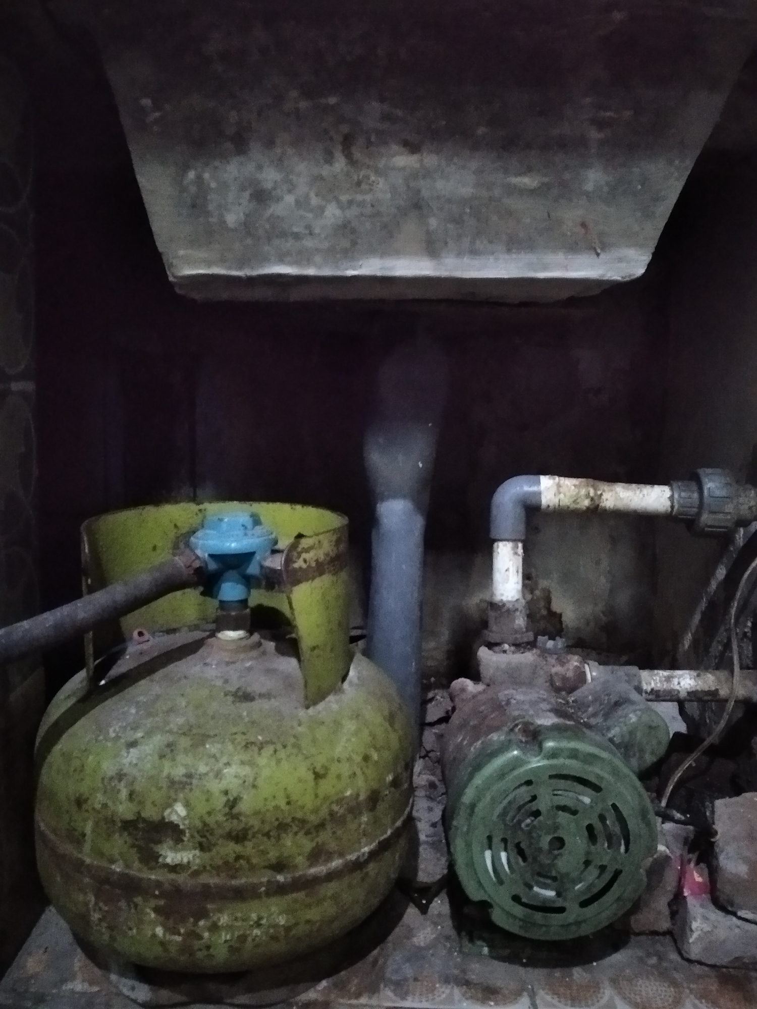 In a dimly lit, enclosed space that appears to be a utility alcove in Rambipuji, Indonesia, a green LPG gas cylinder is positioned to the left. A gray hose connects to the blue regulator atop the cylinder. To the right of the gas cylinder, a green electric pump is visible, with a silver pipe extending from it and connecting to a white pipe angled upwards. The environment is cluttered and appears weathered, with exposed pipes and a rough, possibly concrete, overhead structure. The floor is tiled.