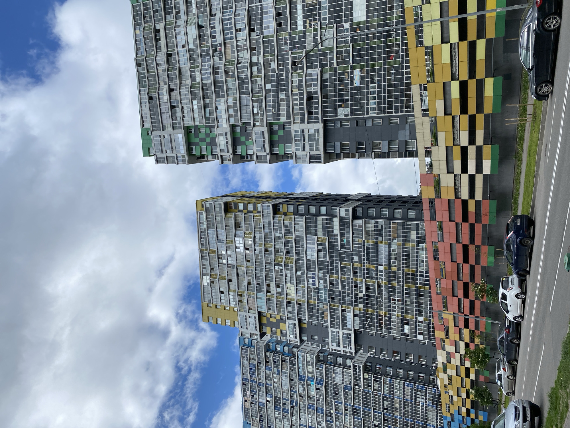 The image captures a scene of modern architecture in an urban environment. Two tall buildings with a dense grid of windows dominate the frame, suggesting residential or commercial apartments. The buildings are distinguished by colorful patterns on their facades; one features yellow, black, green, and white squares, while the other has red, black, green, and white squares. A row of parked cars lines a street running alongside the buildings, indicating an active city setting. The sky, partly visible, displays a mix of blue and white clouds, suggesting a bright or overcast day. Overall, the photograph presents a snapshot of contemporary city life with a focus on architectural design. The location of this scene is in the city of Unknown, Unknown.