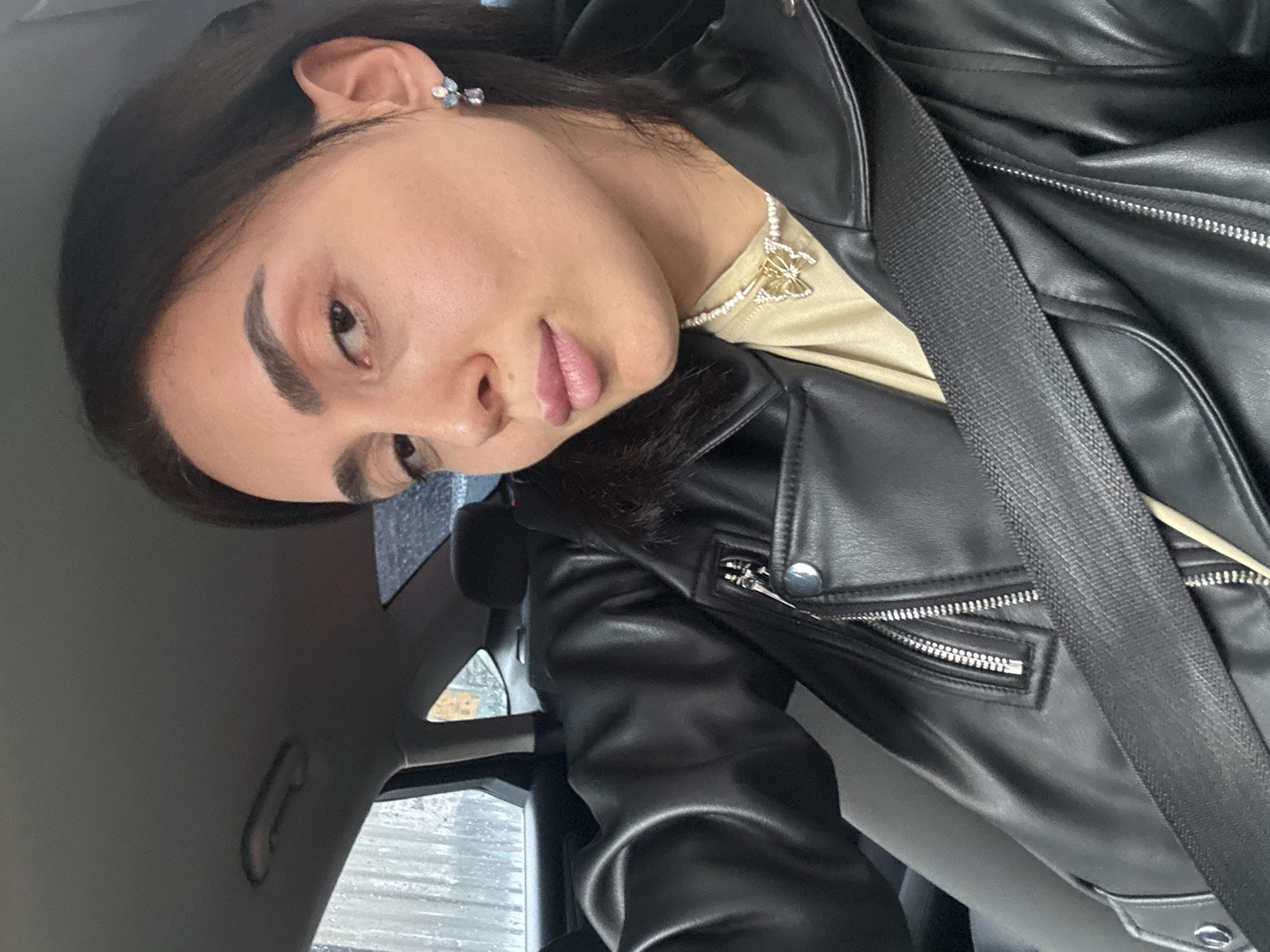 An individual, identified as female, is seated in the rear passenger seat of a vehicle, likely a sedan, in Tbilisi, Georgia. She maintains a direct gaze towards the camera. She wears a black leather jacket featuring silver zippers and snaps, over a light beige top. A white beaded necklace with a golden, gem-studded butterfly pendant is visible, as are small, multi-colored floral-shaped stud earrings in each ear. A black seatbelt is securely positioned across her chest and lap. The vehicle's interior features a grey headliner and dark upholstery. Outside the rear passenger window, water droplets are visible, indicating rain or recent precipitation, against an indistinct urban background.