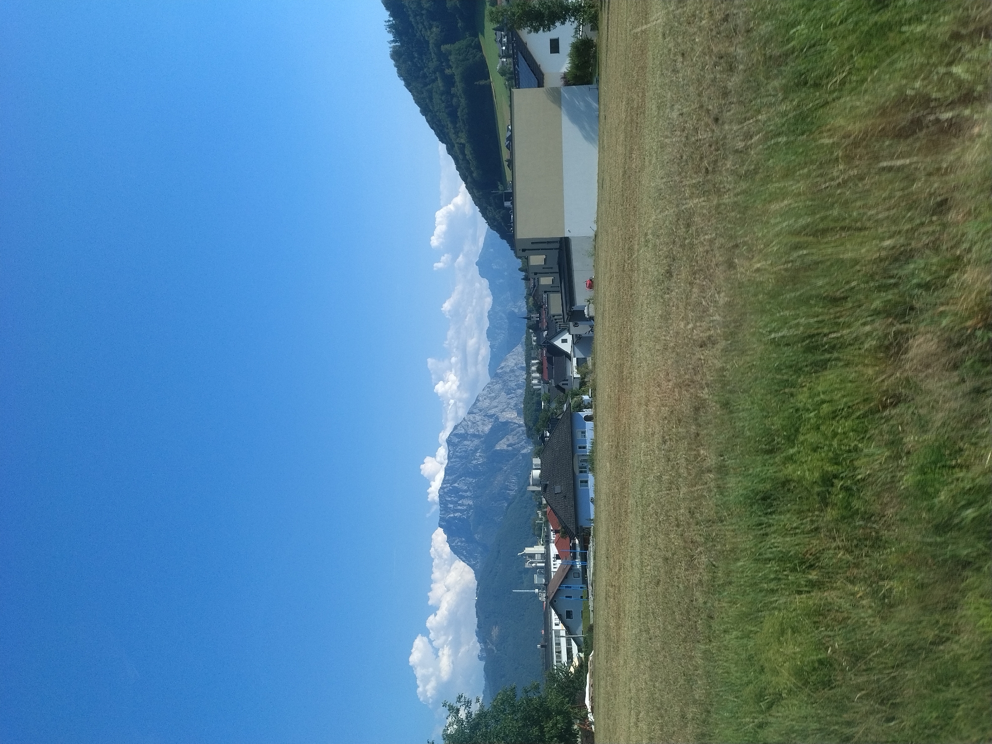 This is a daytime, outdoor, eye-level, medium shot photograph of a rural village in Innergrub, Austria. The foreground is dominated by a field of tall, dry grass, with some patches of greener grass visible. In the middle ground, a cluster of houses and buildings are visible, with a prominent beige and white building on the right side. The background features a large, rugged mountain range under a clear blue sky with scattered white clouds. The overall scene depicts a serene rural landscape with residential structures and natural elements.