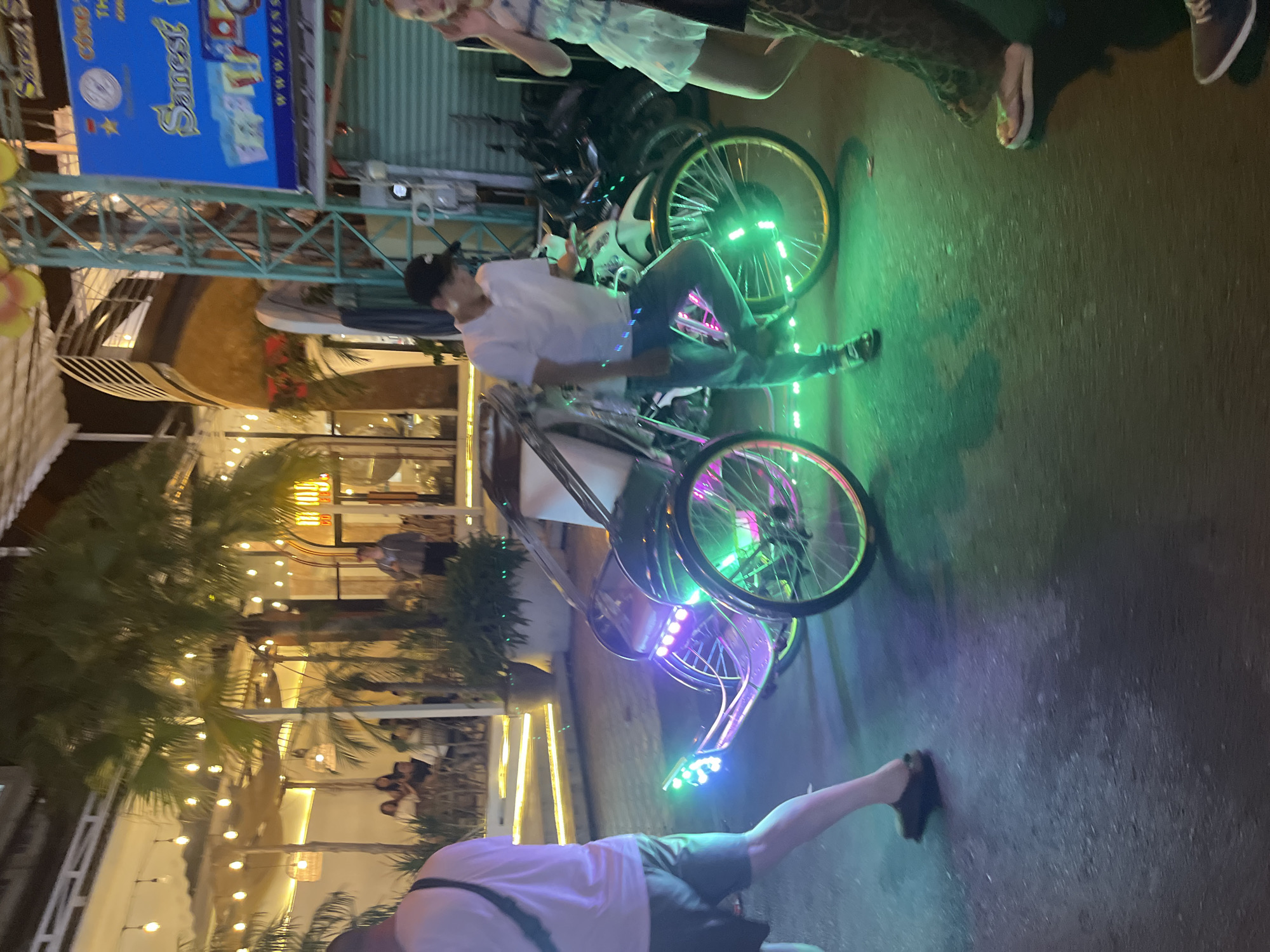 The image captures a street scene at night in Nha Trang, Vietnam, with a focus on a cyclo, a three-wheeled passenger vehicle. The cyclo is adorned with colorful LED lights, primarily green and purple, which illuminate the surrounding asphalt. A young man, wearing a white polo shirt, blue jeans, and a black baseball cap, stands next to the cyclo, appearing to be either operating it or preparing it for a ride. His posture suggests he might be adjusting something on the vehicle or waiting.
In the background, a building with a distinctly curved architectural design is visible, lit by numerous string lights, indicating a commercial establishment, possibly a restaurant or cafe. Palm trees are scattered throughout the scene, their fronds silhouetted against the night sky and the ambient lighting. Further in the background, other people can be seen either inside or near the establishments, but they are not the primary focus.
To the right of the cyclo, a woman in a light-colored dress is partially visible, her presence suggesting other patrons or passersby. The overall atmosphere is lively and illuminated, typical of a tourist-frequented area at night. The ground is wet, reflecting the colorful lights, which could indicate recent rain. The image appears to have been taken from a slightly low angle, emphasizing the cyclo and the surrounding urban environment.