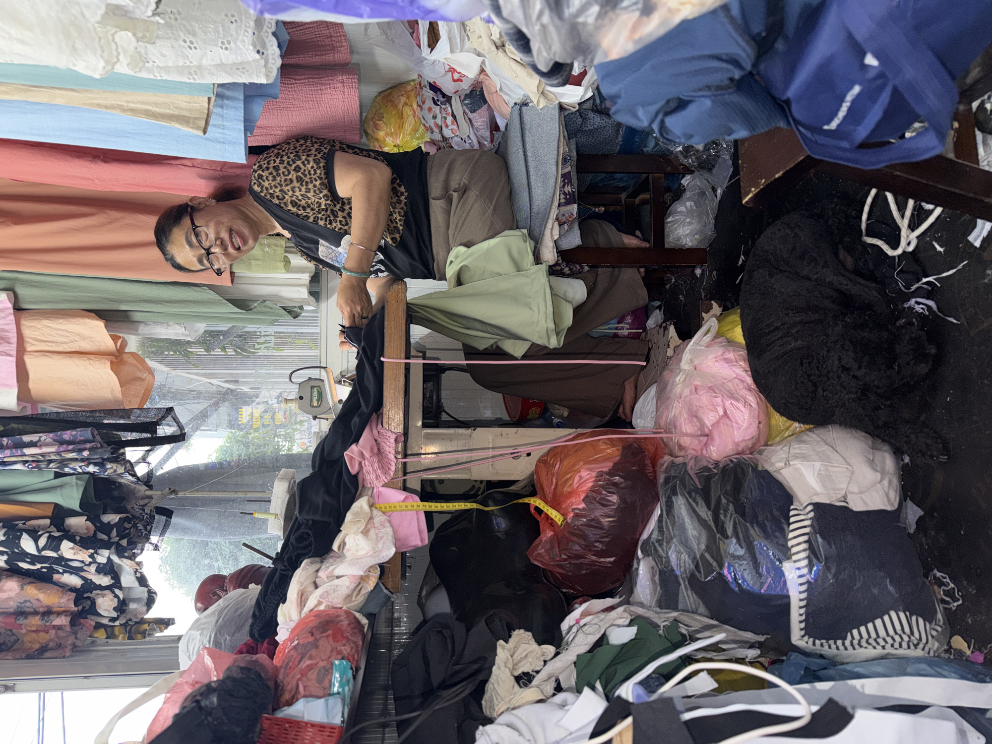 An older Asian woman with glasses is seated in a very cluttered sewing shop, likely in Đà Nẵng, Vietnam. She is wearing a leopard-print top and has her hands near a sewing machine. Fabric of various colors and patterns surrounds her on all sides, piled high around her and obscuring most of the shop's interior. There are also plastic bags filled with clothes. A measuring tape is visible draped around a plastic bag. The shop appears to be very cramped and filled with textiles.