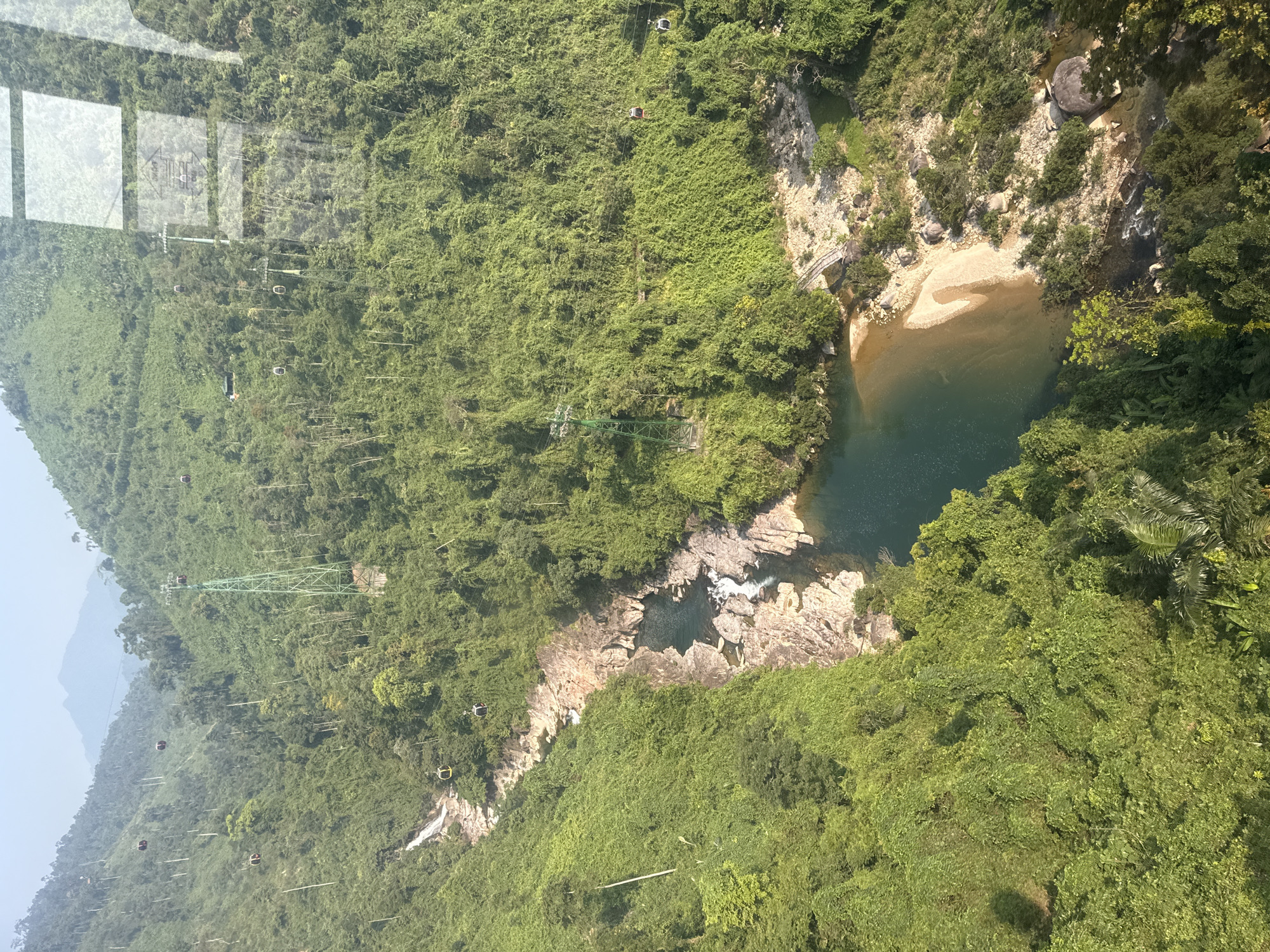 The image depicts a mountainous, forested landscape in Đà Nẵng, Vietnam, viewed from an elevated position, possibly a cable car. A river or stream flows through the scene, forming pools and small waterfalls. Dense green vegetation covers the slopes. A cable car system with support towers and cables is visible extending across the landscape. There are cable cars positioned at intervals along the cable system. Reflections are seen in the upper left corner, indicating a window or enclosure.