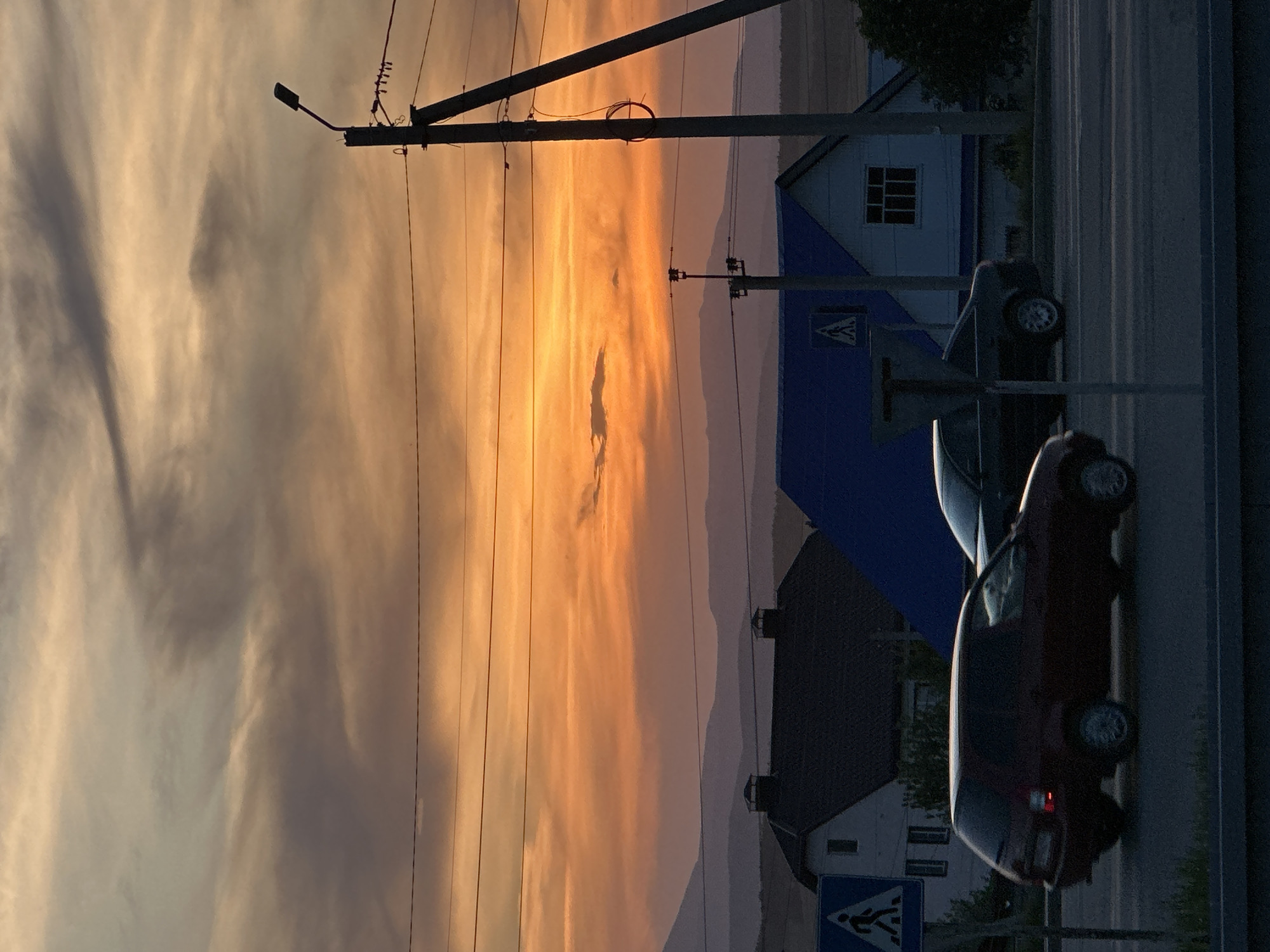 The image shows a street scene during a vibrant sunset, with golden and grey-toned clouds filling the sky. Electrical poles and wires run across the sky, adding a graphic element. On the right side of the image is a road with two dark-colored cars parked or driving on it. Next to the road is a building with a blue and white facade, partially obscured by a pedestrian crossing sign. Behind the building, a range of hills or mountains can be seen in the distance, colored in shades of pink and purple by the setting sun. A house with a dark roof and dormer windows is visible further back. The location appears to be a roadside or residential area within the city of Unknown, Unknown.