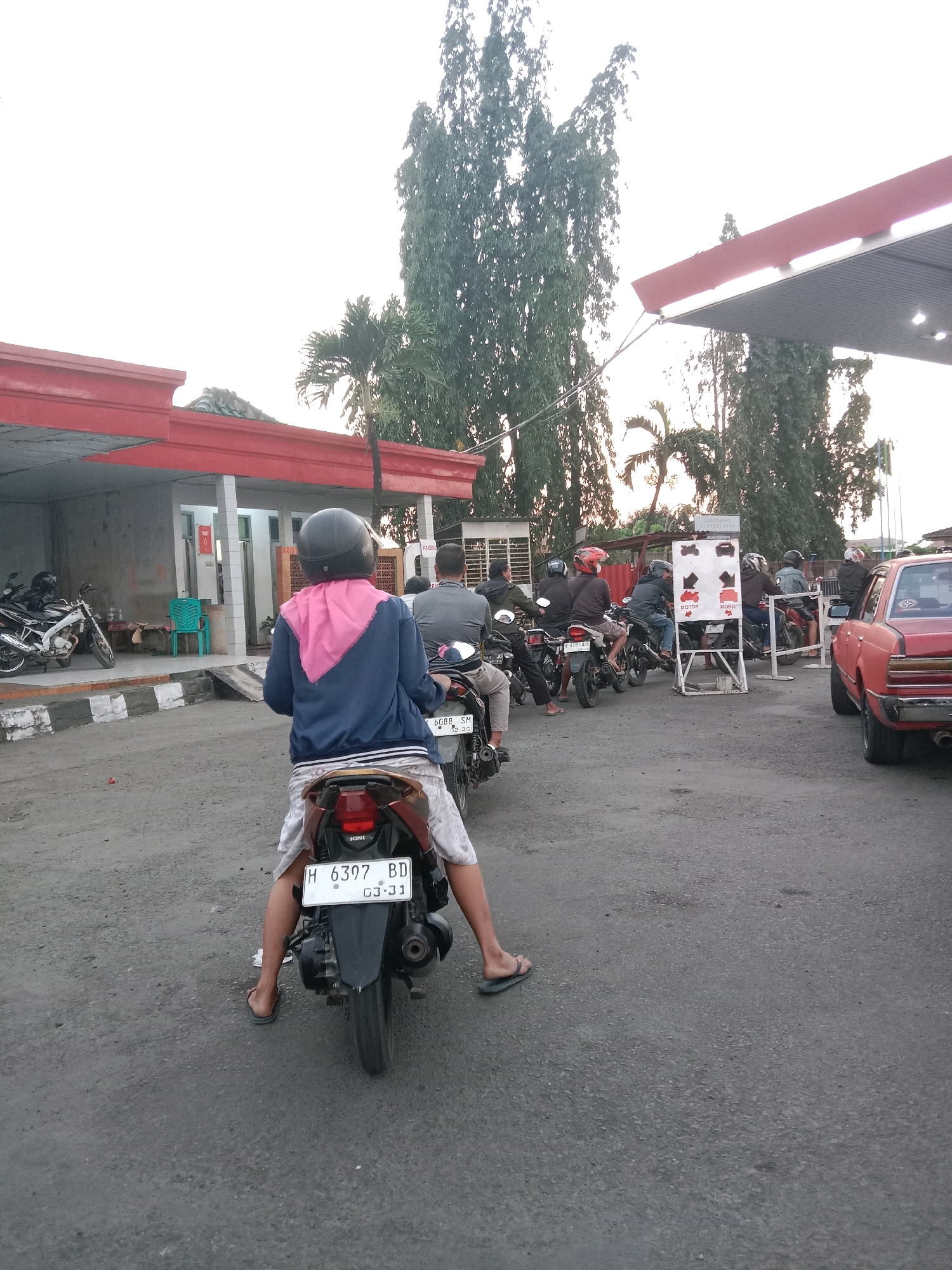 The image depicts a street scene in Brangsong, Indonesia, likely at a gas station. In the foreground, a person wearing a helmet and a pink scarf over a blue jacket is seated on a dark-colored motorcycle. The motorcycle has a license plate that reads "H 6397 BD 03-31". The person's legs are bare, and they are wearing flip-flops. The motorcycle's license plate is clearly visible, indicating it's a vehicle in Indonesia.
Behind this motorcycle, several other motorcycles and their riders are visible, forming a queue. Some riders are wearing helmets, while others are not. One motorcycle has a license plate that reads "L 4688 SM".
In the background, a red-and-white structure, possibly a part of the gas station canopy, is visible. Tall trees, including palm trees, are scattered throughout the scene, suggesting a tropical or subtropical climate. A red car is parked on the right side of the image. A white sign with red and black icons, possibly indicating different types of vehicles, is also present. The ground is paved with asphalt. The overall atmosphere appears to be that of a typical day in a moderately busy Indonesian town.