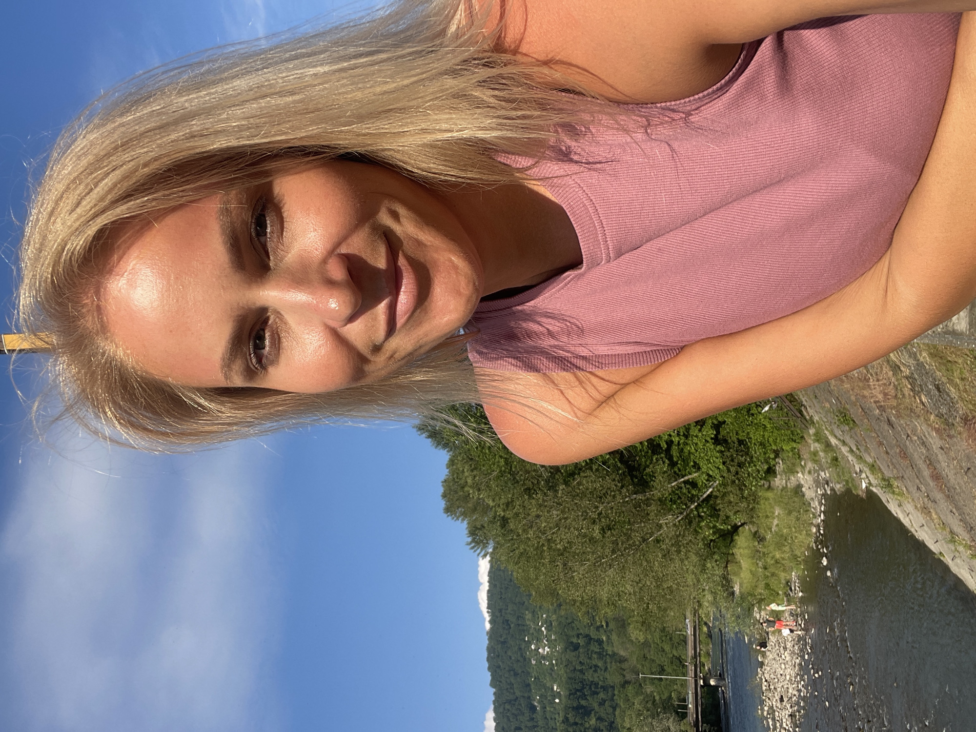 A blonde woman smiles into the camera, positioned on the right side of the frame. She is wearing a pink, ribbed tank top and appears to be outdoors. Behind her, a body of water, likely a river, flows with a rocky shore. A bridge spans the river in the distance. Trees and a lush, green mountainside with scattered houses are visible on the left side of the image, with a bright blue sky above. This scenery is characteristic of the natural beauty found in and around Sochi, Russia, a city known for its Black Sea coast and mountainous terrain.