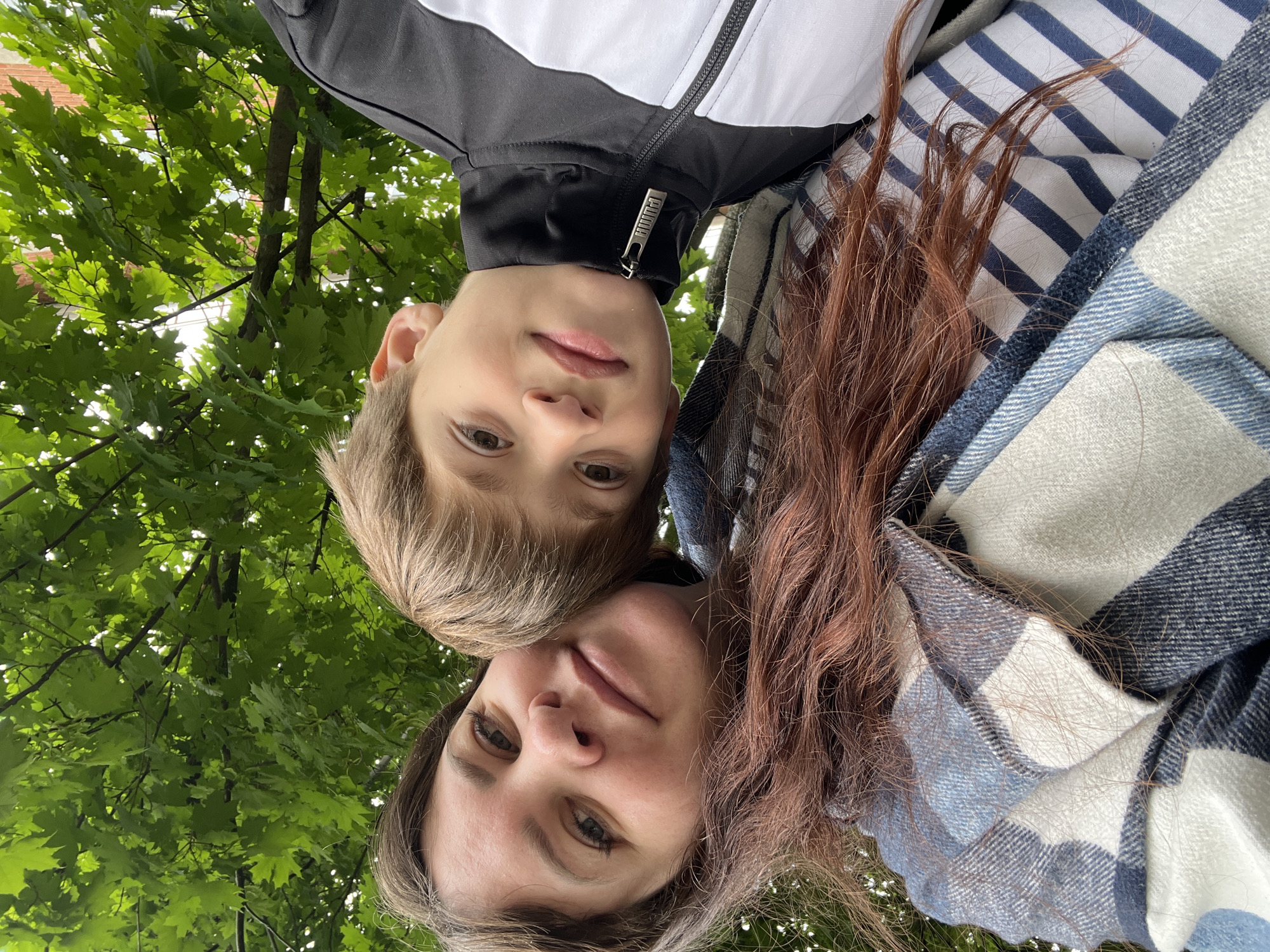 This media file presents a unique, inverted perspective of an adult woman and a child, likely a mother and son, looking down towards the camera. Their faces appear upside down in the frame, set against a backdrop of vibrant green tree foliage and bright sky.
The woman, positioned towards the bottom of the frame, has long, reddish-brown hair and is wearing a blue, white, and light-grey plaid item, possibly a shirt or a blanket wrapped around her. Her expression is calm, with a subtle smile.
Above her, the child, with light brown hair, wears a black and white athletic jacket. A metallic zipper pull on his jacket is clearly visible, featuring the "PUMA" brand logo. His expression is neutral and direct.
The setting is outdoors, beneath a canopy of dense green leaves and branches, through which patches of bright sky are visible. The low-angle shot suggests the photo was taken from directly below the subjects, looking up at them and the natural surroundings. The bright natural lighting indicates it is daytime. The overall mood conveyed is one of candidness and tranquility, possibly during leisure time spent in a park or wooded area in Noviy, Russia.