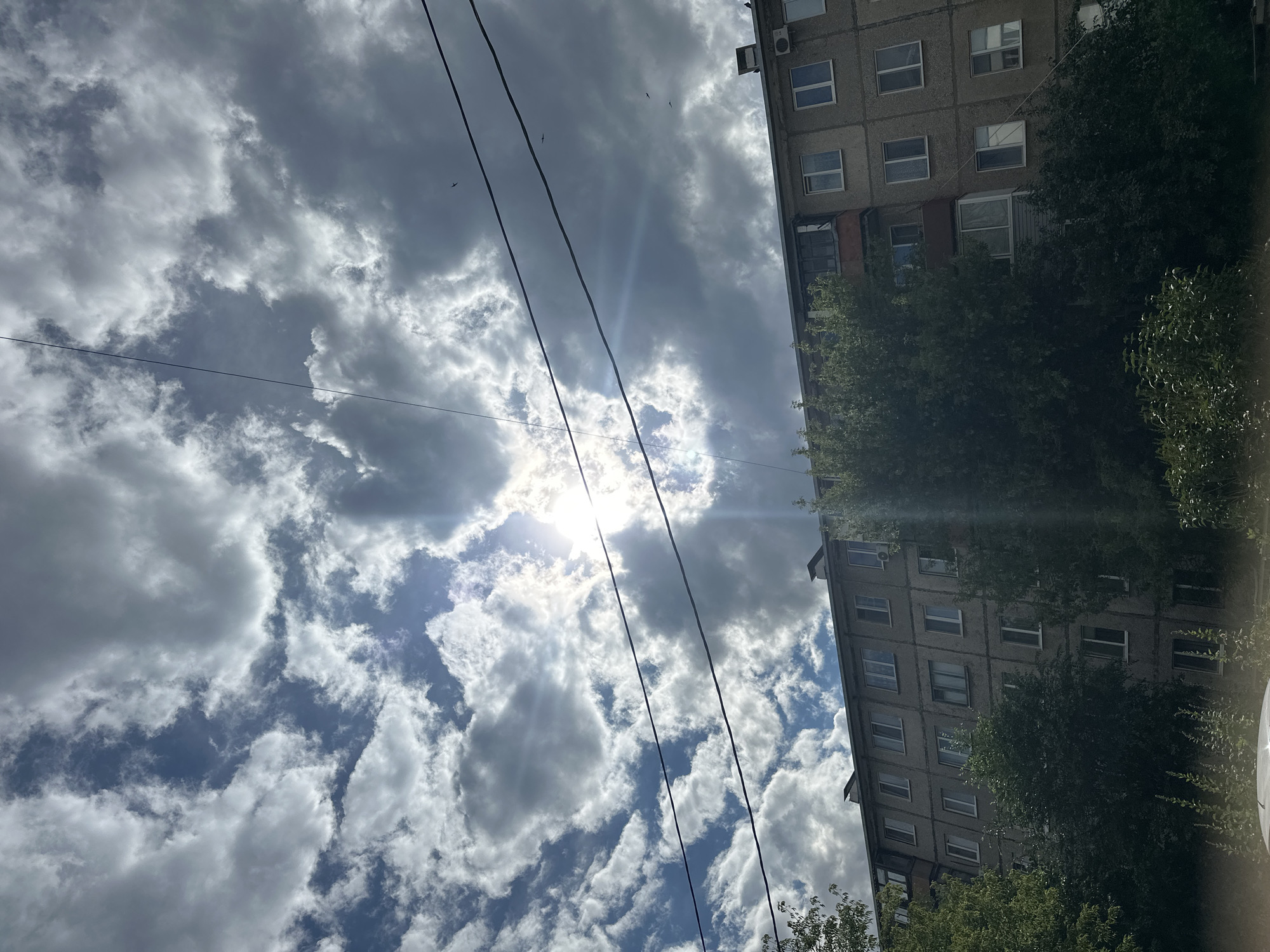 An eye-level outdoor shot captures a cloudy sky above a drab, concrete apartment building in Orsk, Russia. The sun attempts to break through the clouds, creating a bright glare and lens flare in the upper center of the frame. Two black electrical wires stretch horizontally across the sky. The apartment building is typical of Soviet-era architecture, with numerous windows and balconies. Lush green trees and foliage obscure the lower portion of the building and the foreground. The scene conveys a sense of urban normalcy, likely during daytime.