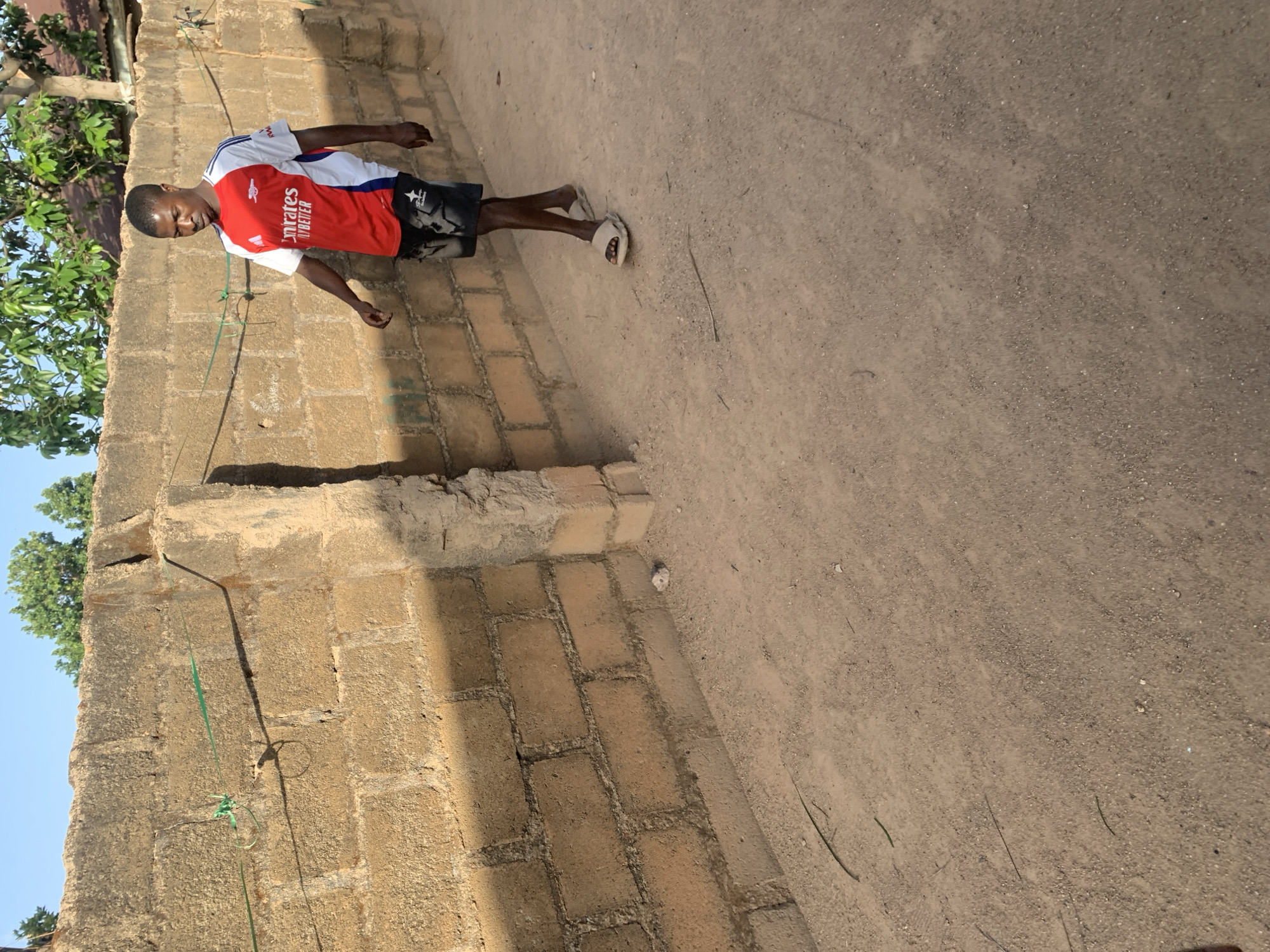 A man stands outdoors between a brick wall and a dirt-covered area. The man is wearing a red and white soccer jersey, dark shorts, and sandals. The brick wall is constructed of light brown bricks with visible mortar. A green string hangs down the wall. The dirt-covered area is light brown and covers a large portion of the image. The location is Jos, Nigeria.