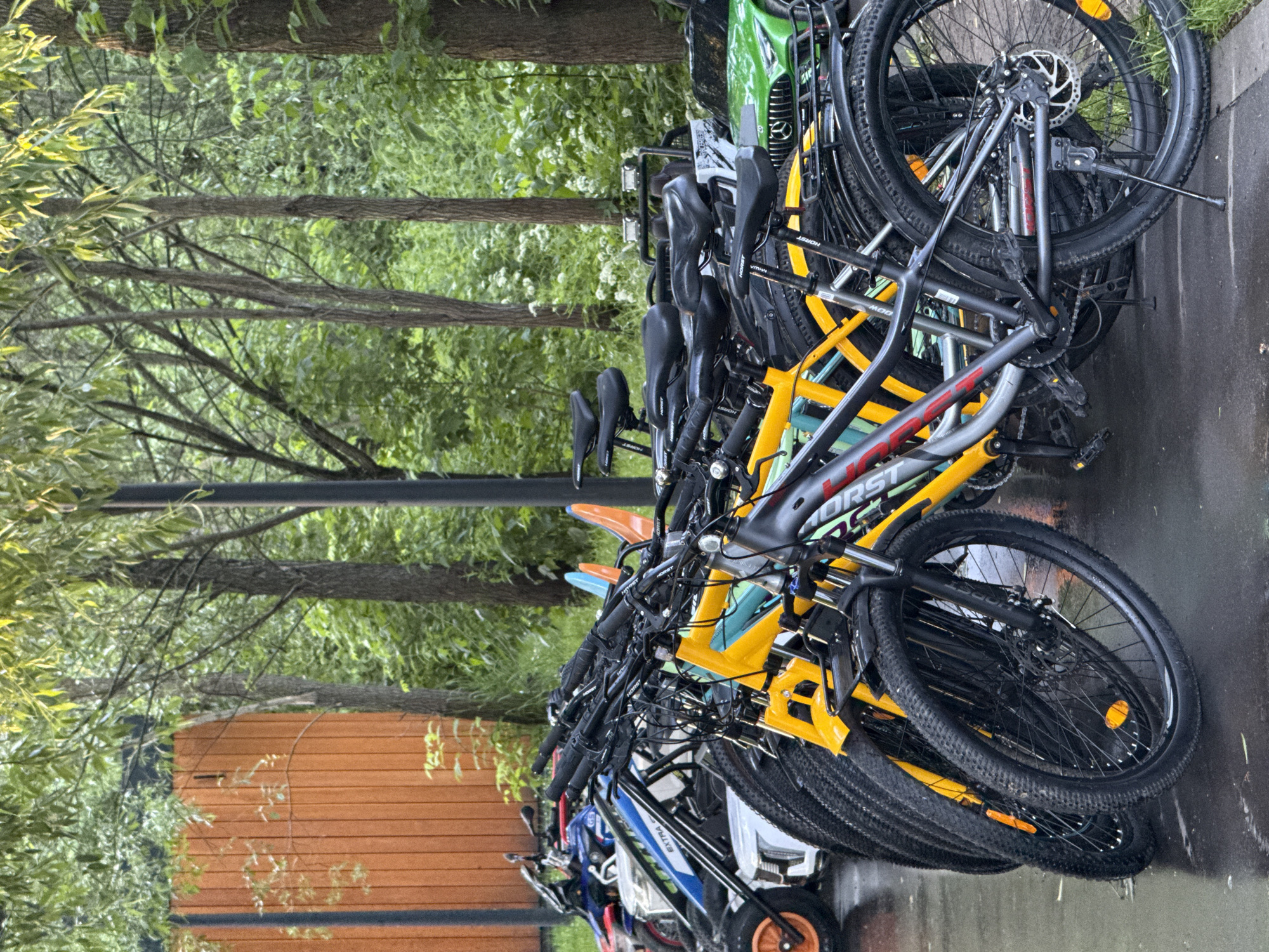 A row of bicycles and electric scooters are parked on a paved surface in front of a wooden fence and a lush green forest. The bicycles are of various colors, including yellow, blue, and gray, and some appear to be electric. Some of the bicycles have lights mounted on their handlebars. In the background, several trees with dense foliage are visible. A dark gray pole runs vertically through the center of the frame. The ground is wet, suggesting recent rain. The scene appears to be a public area, possibly a park or rental station, in Moscow, Russia. There are no people visible in the image.