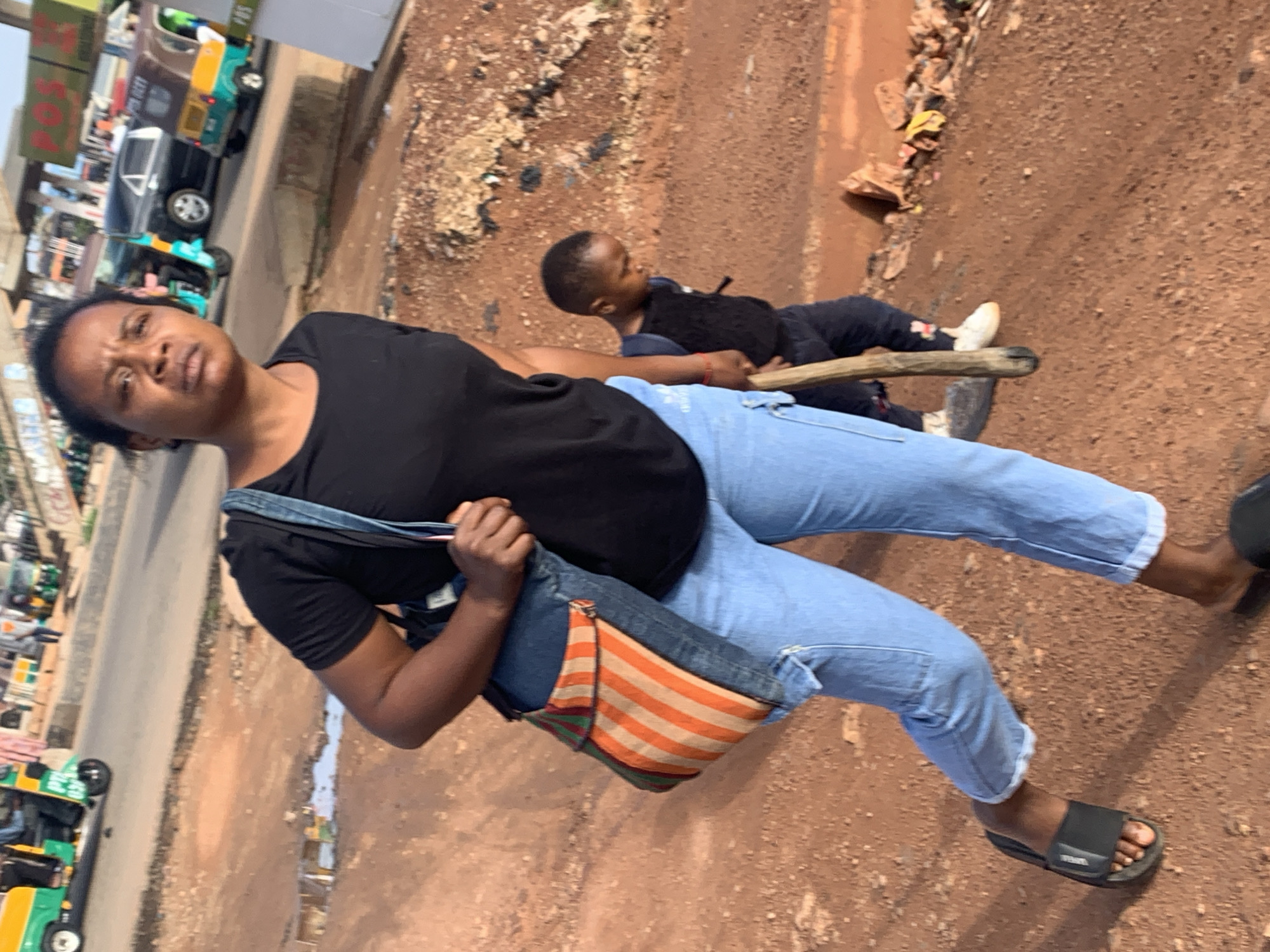 In Gura Top, Nigeria, a Black woman in a black t-shirt and light blue jeans walks on a dirt path. She is carrying a patterned shoulder bag. Behind her, a Black child in a dark shirt and pants walks with a wooden stick. In the background, there are several vehicles including a green and yellow auto-rickshaw, a dark car, and another colorful auto-rickshaw. The ground is reddish-brown dirt, with some puddles of water and scattered debris. A busy street with more vehicles and buildings is visible in the distance.