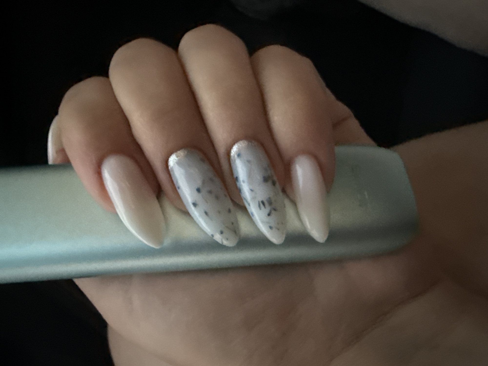 A close-up shot shows a person's hand with manicured nails holding a light blue cylindrical object. The nails are long and almond-shaped. Some nails are painted a milky white, while others feature a marbled pattern of white and gray with small blue flecks, and a subtle silver glitter detail at the tips. The background is dark and out of focus. The image is shot from a high angle, looking down at the hand and object. There is no discernible location in Burgas, Bulgaria, or any other specific geographical context.