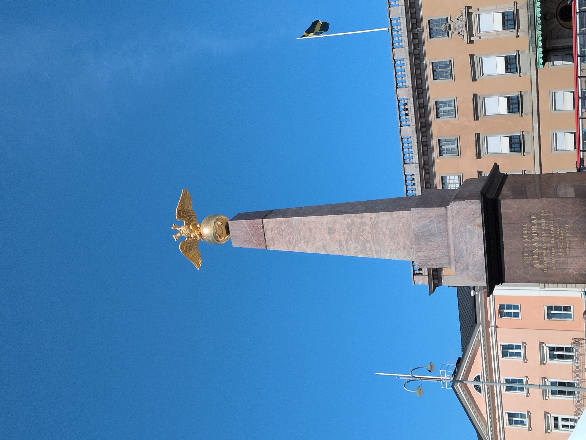 The image shows the Alexander Nevsky Monument in Senate Square, Helsinki, Finland. The monument is an obelisk topped with a golden double-headed eagle clutching a globe. The obelisk is made of red granite and has an inscription in Latin. In the background, there are several buildings, including the Helsinki Cathedral and the Government Palace. A Finnish flag is visible on a flagpole. The sky is clear and blue. There are no people visible in the image.