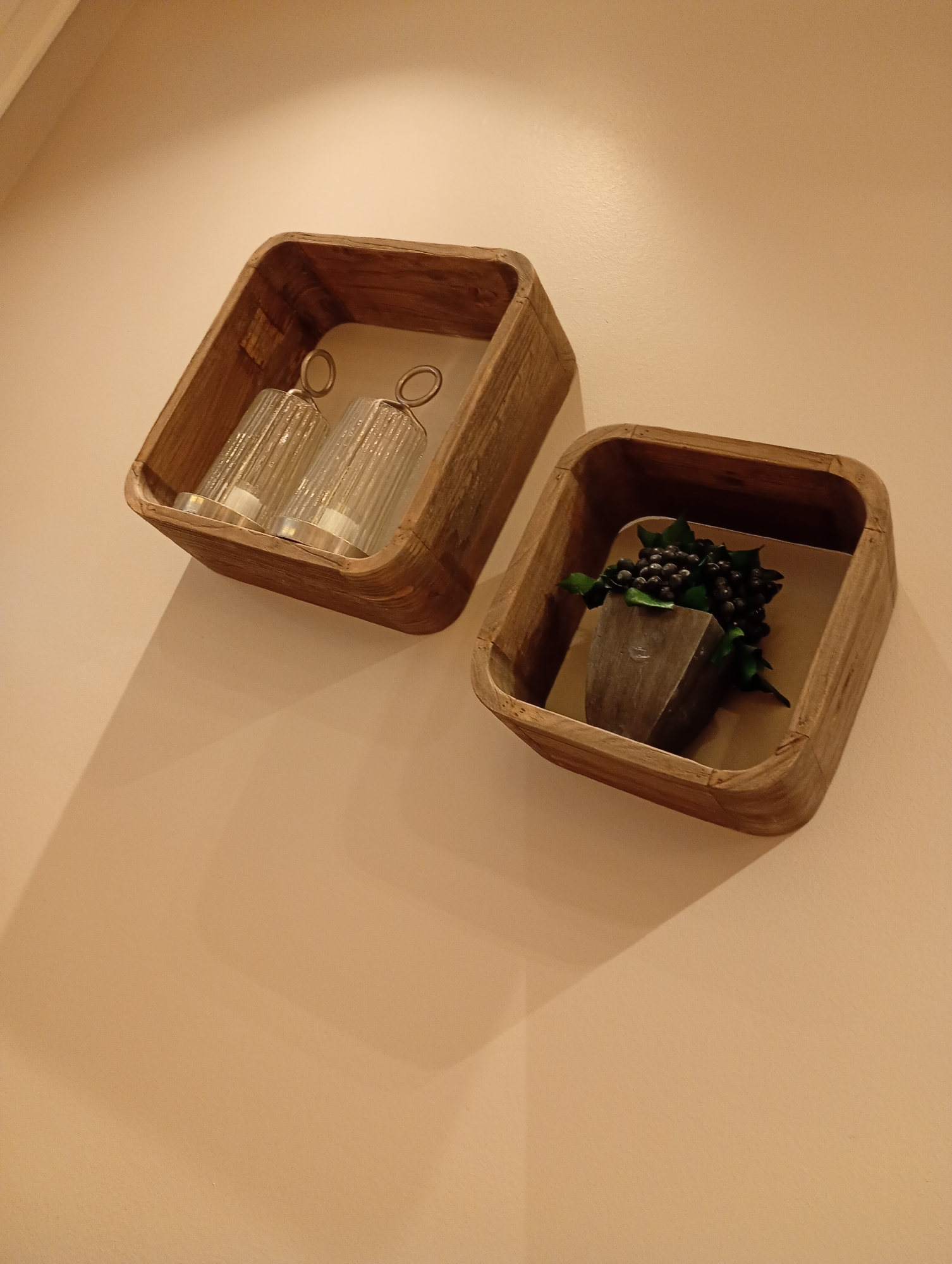 Two wooden, open-faced wall shelves are mounted on a light beige wall. The shelf on the left contains two clear glass cylindrical objects with silver tops and looped handles, possibly candle holders or decorative jars. The shelf on the right holds a dark, triangular object from which a cluster of dark berries and green leaves emerges, likely a decorative arrangement. The lighting casts shadows from the shelves onto the wall. The image was taken at the "Haus Am Sonnenberg" in Bad Sachsa, Germany.