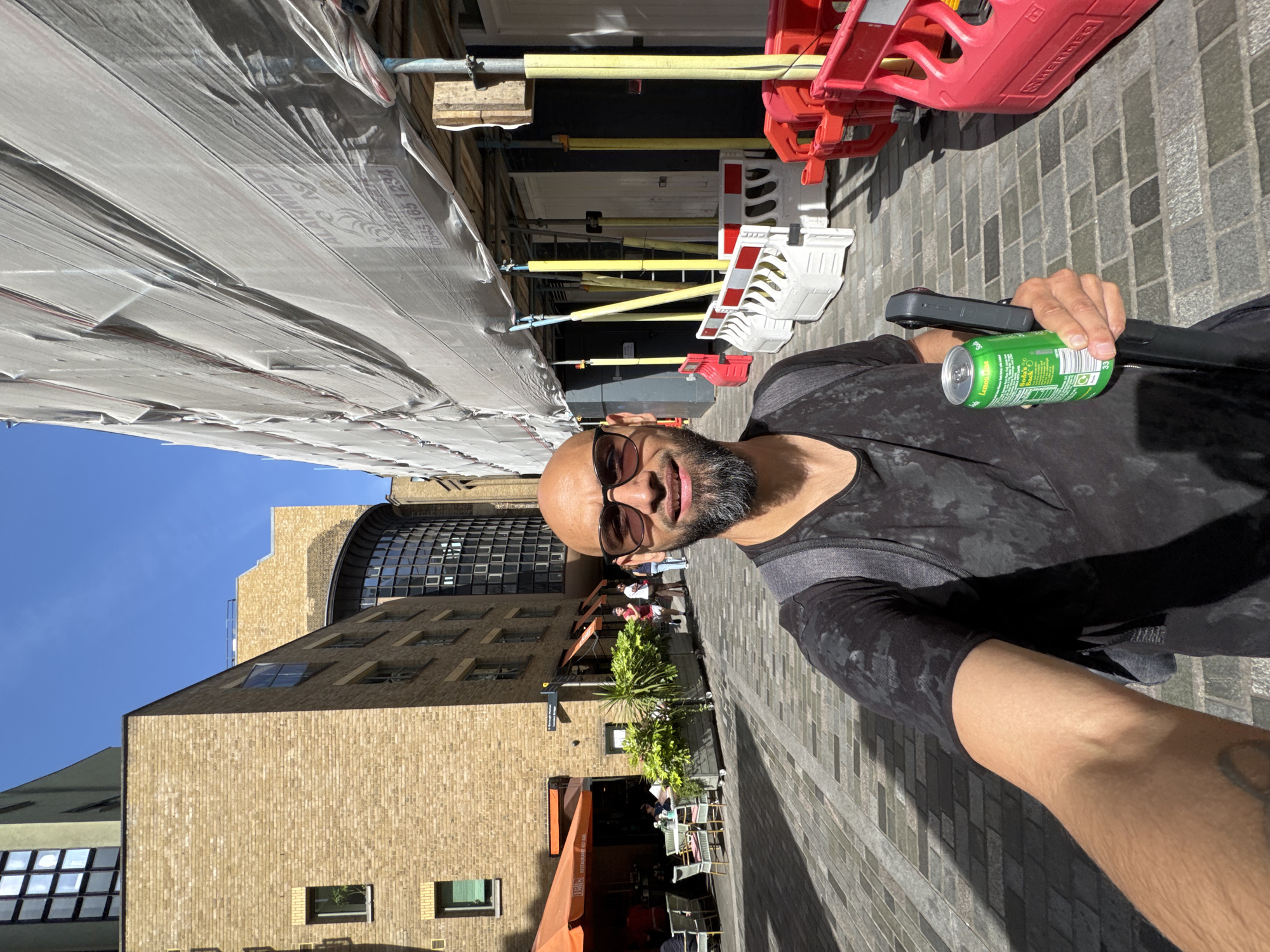 The image shows a person holding a green can of soda and a black object, possibly a phone grip or a selfie stick, in a pedestrian area. The person has a shaved head, dark sunglasses, and a beard. The background features a brick building and a construction site.
Location: The scene is likely in the city of London, United Kingdom, based on the architectural style of the brick building, the paving, and the presence of a construction site. A restaurant called Bill's Restaurant & Bar is seen in the image. The overall environment and architectural elements are characteristic of London.
