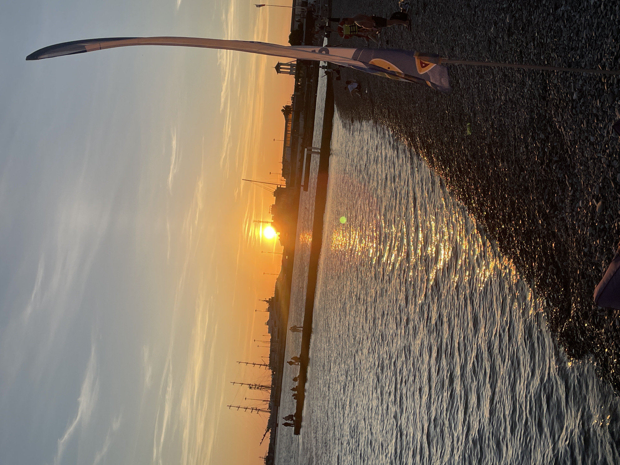 This media file depicts a serene sunset scene at a pebbled beach, likely in Sochi, Russia. The sun, a bright orb, is low on the horizon, casting a warm, golden glow across the sky and the rippling surface of the water. Wispy clouds are scattered across the sky, tinged with orange and yellow from the setting sun.
In the foreground, a tall, curved object, possibly a streetlamp or a decorative structure, stretches into the frame from the left, its dark silhouette contrasting with the bright sky. A sliver of a blue and white patterned flag is also visible, attached to a thin pole extending from the top right of the frame. The beach itself is covered in dark pebbles, reflecting the sunlight.
Along the waterline, several silhouetted figures are visible, suggesting people enjoying the evening. Some appear to be walking or standing along a pier or boardwalk that extends into the water. In the distance, the outline of buildings and what appear to be tall ships with masts are visible against the sunset. The overall mood is peaceful and tranquil, capturing the beauty of a coastal sunset. There is no visible text in the image.