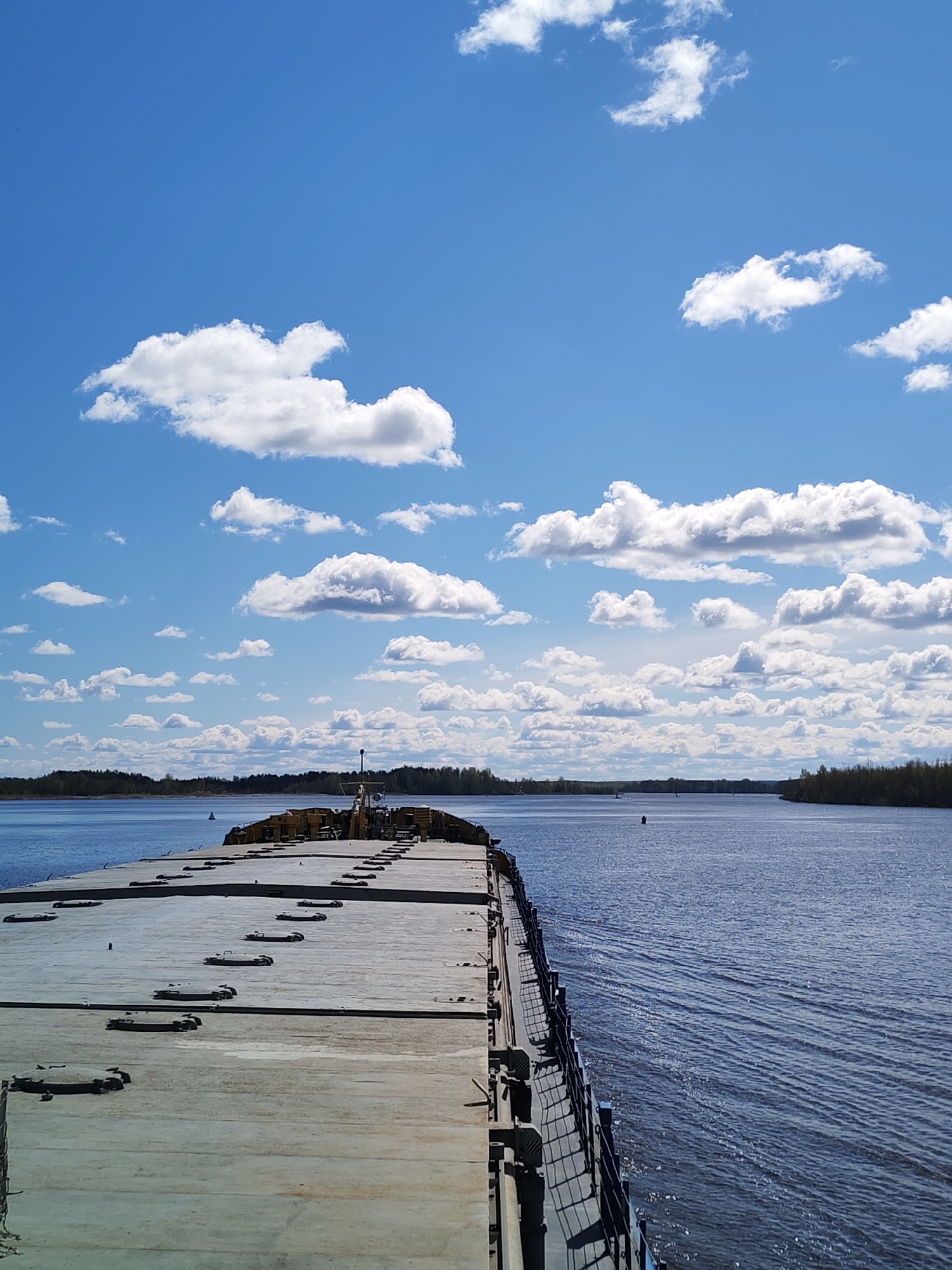The image depicts a barge on a wide river near Kargulino, Russia. The barge has a flat, concrete surface with hatches and railings along the edge. The river is wide and calm, with trees visible on both shores. The sky is blue with puffy white clouds. A small buoy is visible in the water, as is another boat on the horizon.