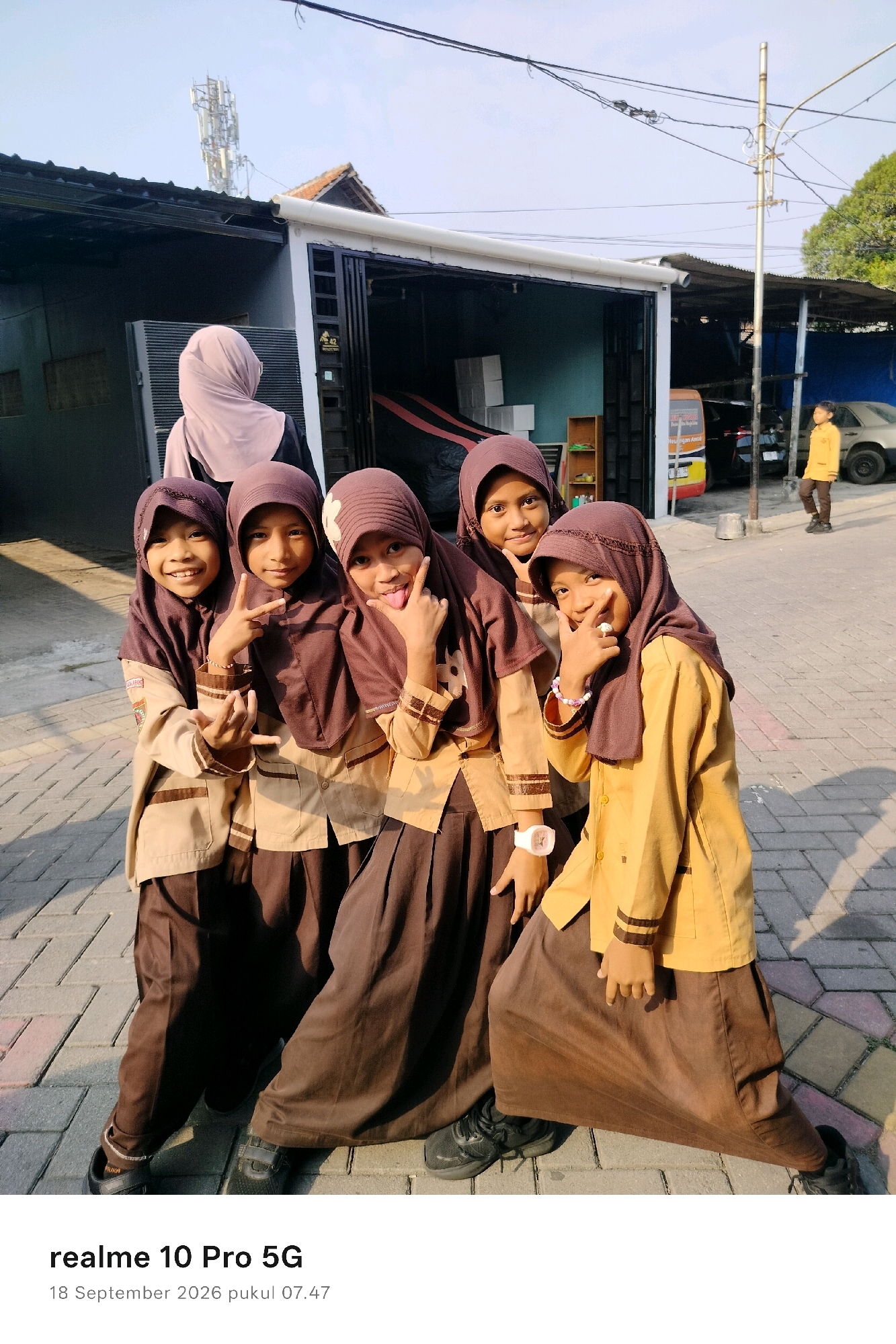 In Semarang, Indonesia, five young girls in school uniforms, consisting of brown skirts and light brown shirts with dark brown hijabs, pose for a photo. The girl on the far left smiles broadly, making a gesture with her hand. The second girl from the left makes a peace sign with her fingers. The middle girl sticks out her tongue playfully. The second girl from the right makes a hand gesture with her thumb and index finger near her mouth. The girl on the far right is also posing. They are standing on a paved walkway in front of a building with a dark metal facade and a corrugated metal roof. In the background, a cell tower is visible against a clear sky, along with trees and a parked car. The image captures a moment of youthful camaraderie outdoors.
