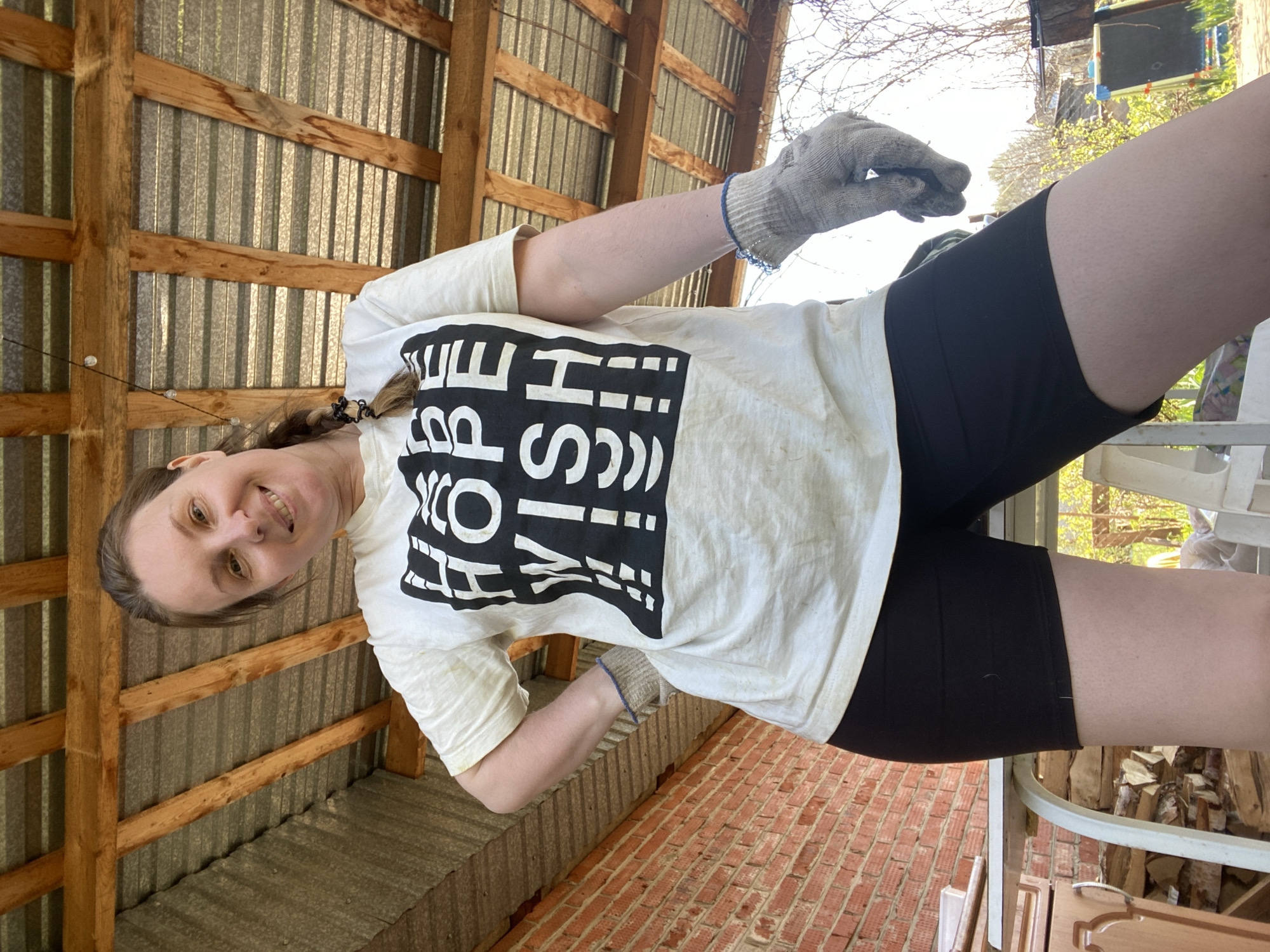 A person, possibly female, is wearing a white t-shirt with a large black square design that includes the words "HI! HELLO! YOU'RE AWESOME" in a stylized arrangement. The person is also wearing black shorts and work gloves. They are standing outside, possibly in a rural area, with a brick wall and a corrugated metal structure visible in the background. The location is not explicitly stated as Chaikovsky, Russia, in the image, and there is no definitive evidence to confirm that. The person is smiling and appears to be looking at the camera. They are holding something in their gloved hand, but the object is not clearly visible.
