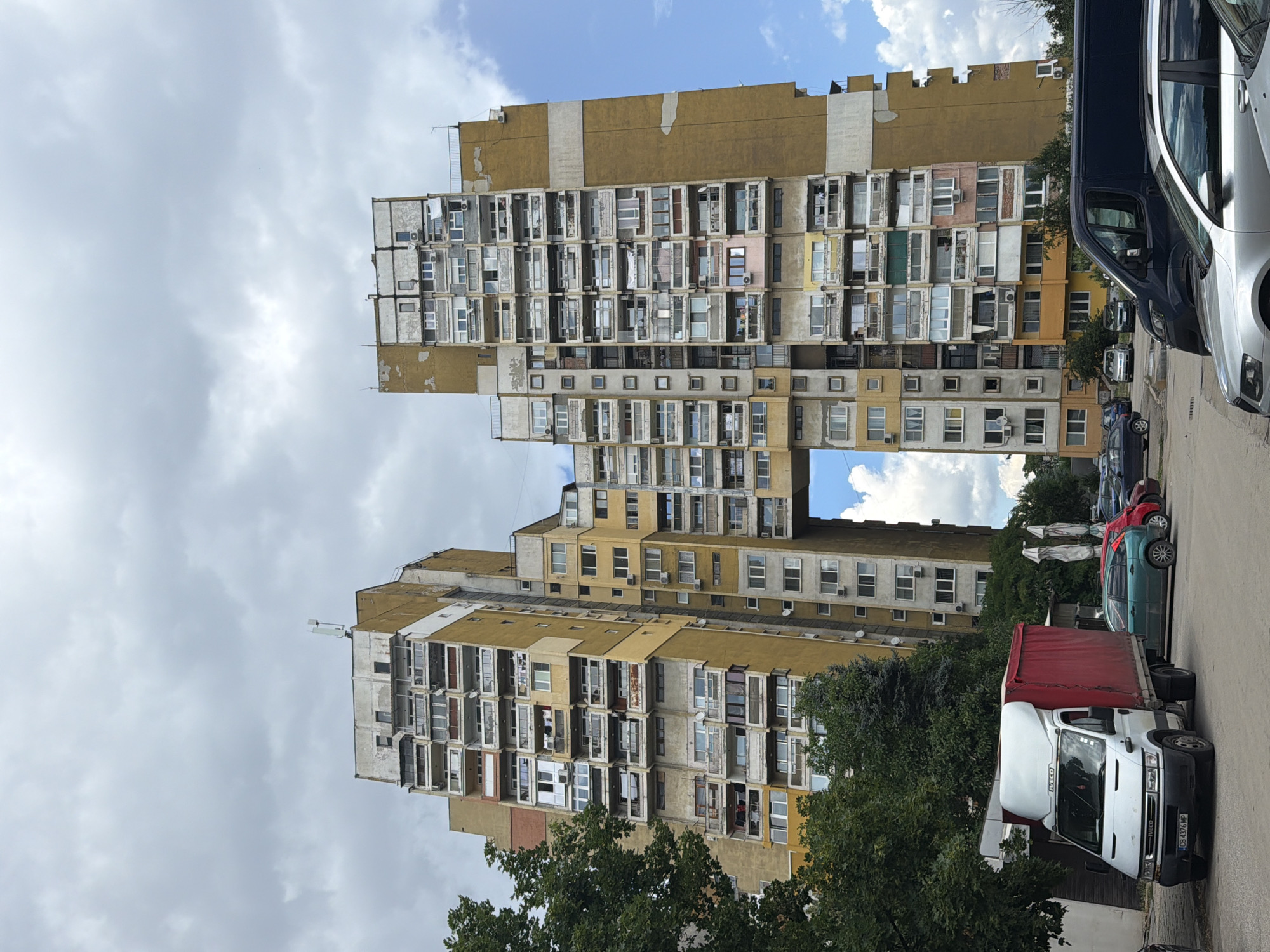 This is a vertical, full-shot photograph of an apartment building in Sofia, Bulgaria. The building is an unusually shaped, Brutalist structure with a distinctive H-shape. It is predominantly yellow and grey, with numerous balconies and windows. The building appears to be in a state of disrepair, with some visible wear and tear.
In the foreground, to the right of the building, there is a street with several vehicles parked. These include a white truck with a red tarped cargo bed, a dark blue van, a silver car, a green car, and a red car. The truck has "IVECO" printed on the front. The license plate of the white truck reads "CB 4376 MP". The sky is cloudy with patches of blue.
The scene suggests a typical urban environment in Sofia, likely during the daytime. The weather appears to be partly cloudy. The overall impression is one of a lived-in, slightly aged, but still functional urban landscape. There are no people clearly visible in the image.