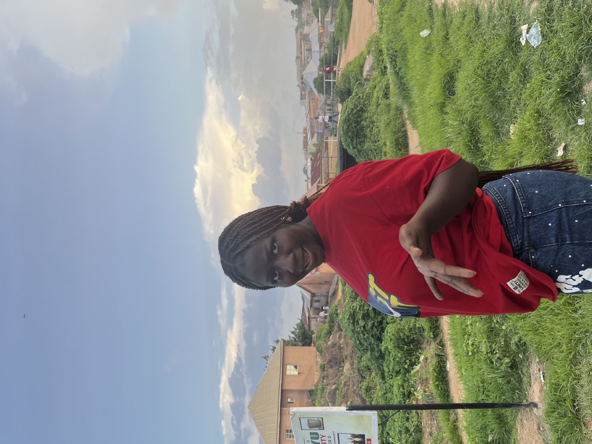 A young Black woman with braided hair and wearing a red t-shirt and blue jeans stands on a grassy embankment, looking at the camera with a playful expression and her hand held up. In the background, a signpost is visible on the left, and beyond it, a cluster of buildings, including a house with a beige roof, are situated on a hill. Further in the distance, more buildings and a bridge are visible, suggesting an urban or semi-urban setting. The sky above is partly cloudy with hints of sunlight breaking through. The overall scene appears to be outdoors in the city of Jos, Nigeria, with a focus on the woman as the central subject.