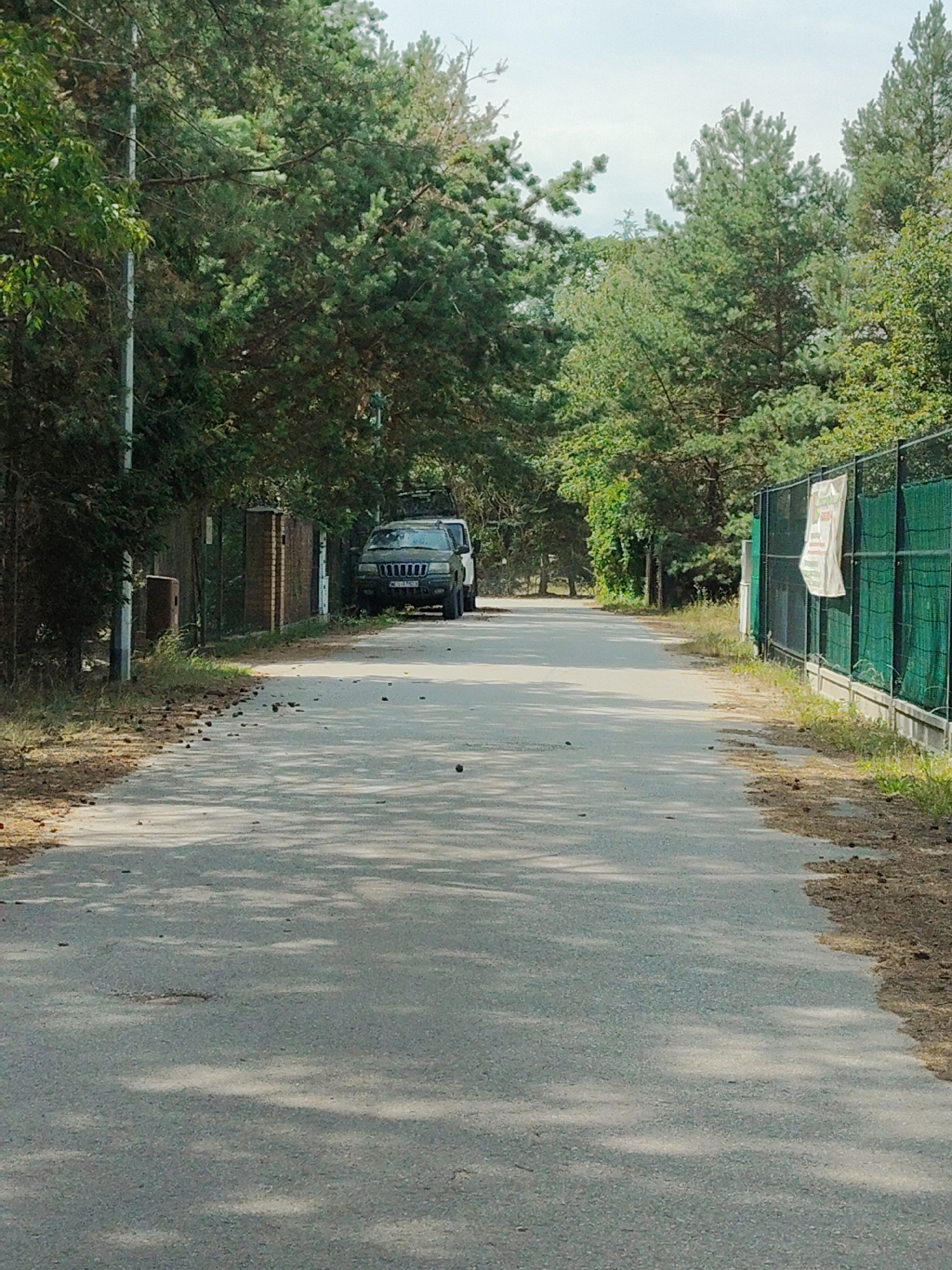 A medium shot captures a paved road lined with trees and fences on both sides, leading towards a parked dark green SUV. The scene appears to be in a wooded area, with tall pine trees and other greenery flanking the road and the fences. On the left side of the road, a dark brown, cylindrical object, possibly a waste bin, stands near a fence made of dark bricks and metal posts. A tall, thin pole with a light fixture is also visible on the left.
On the right side of the road, a dark green metal fence with a net-like covering extends into the distance, with a white banner hanging from it. The banner has some faded red and green lettering, but it is not legible. The SUV, a Jeep Grand Cherokee, is parked on the right side of the road, partially obstructing the view further down the road. The vehicle has a license plate that appears to have alphanumeric characters.
The road surface is asphalt and shows signs of wear and tear, with some debris scattered on it, including what appear to be pinecones and small stones. Patches of sunlight and shadow are cast across the road, suggesting it is daytime with intermittent cloud cover. The overall atmosphere is peaceful and rural, with no people visible in the frame. The presence of trees and the natural ground cover hints at an outdoor setting, likely a residential area or a park, in Palmiry, Poland. The weather appears to be fair, with a bright sky peeking through the trees.