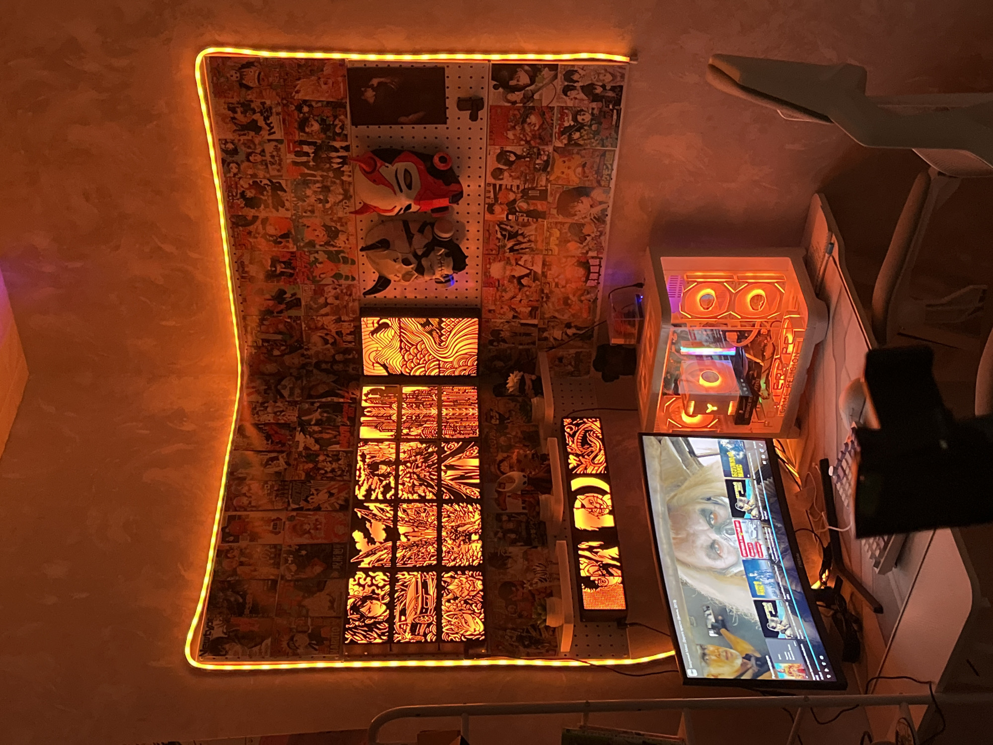 The image depicts a home office or gaming setup with an orange aesthetic. The walls are adorned with numerous anime posters and framed art pieces, illuminated by a vibrant orange LED light strip that outlines the corner of the room. Below the posters, there are two shelves displaying various anime figurines and decorative masks, including what appears to be a Japanese oni mask and a red and white mask.
In the center of the setup is a desk with a widescreen monitor displaying a movie scene. To the right of the monitor is a white computer tower with glowing orange internal lights, featuring a transparent side panel. A keyboard and mouse are positioned in front of the monitor. A white ergonomic chair is partially visible to the right of the desk. The overall environment suggests a personalized and decorated space, possibly for entertainment or gaming. The image does not contain any explicit information to determine its location within Moscow, Russia.