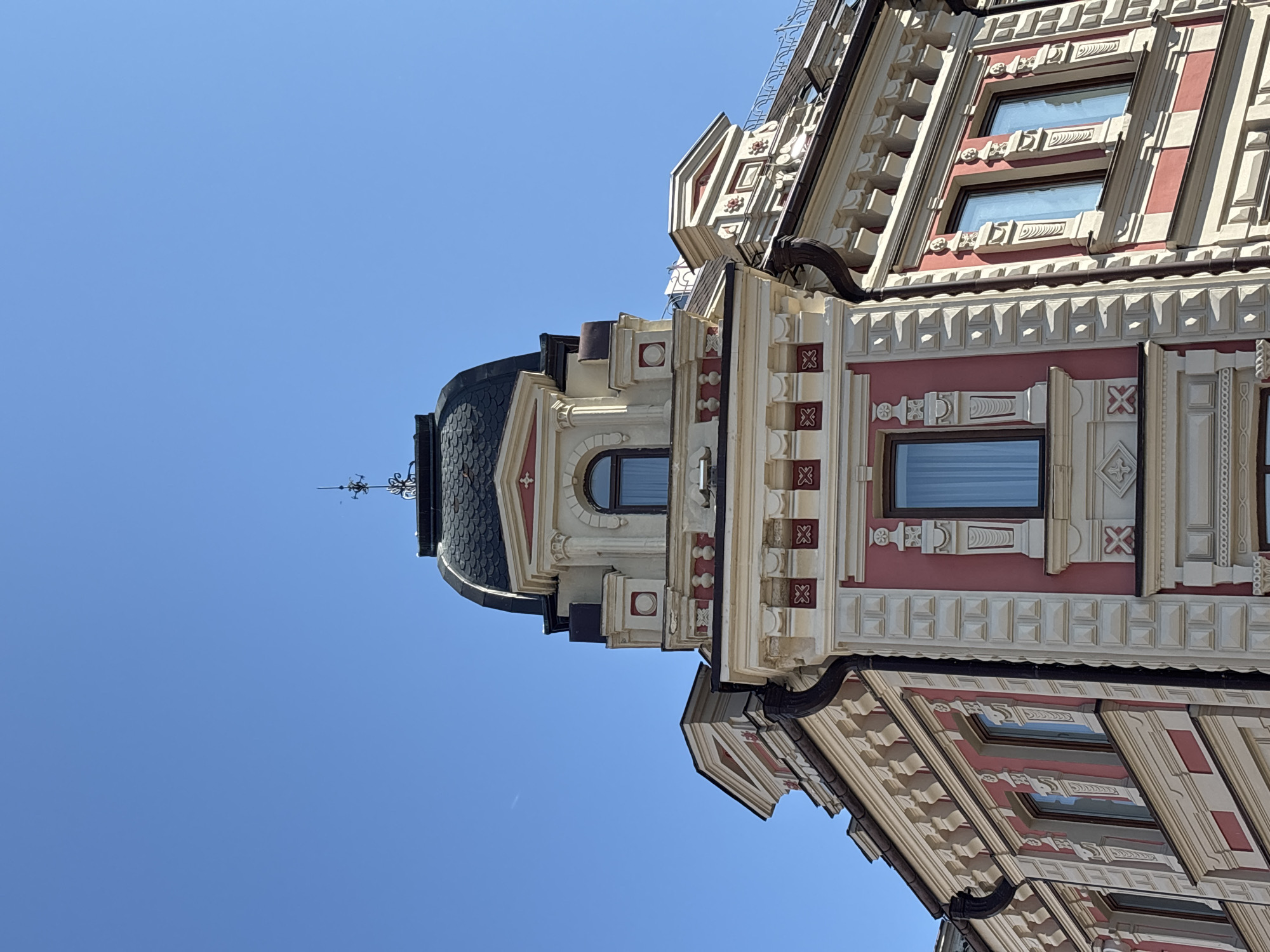 The image shows the ornate facade of a historic building under a clear blue sky. The architecture features a domed roof topped with a weather vane, decorative cornices, and recessed windows with arched detailing. The building's exterior is painted in a combination of cream and pink, with intricate white stucco work and repeating geometric patterns. The corner of the building is visible, suggesting it is part of a larger structure. The architectural style is reminiscent of late 19th or early 20th-century European buildings. This building is located at 59 Karl Marx Street in Kazan, Russia, a city known for its rich history and diverse architectural heritage. No people are visible in the image.