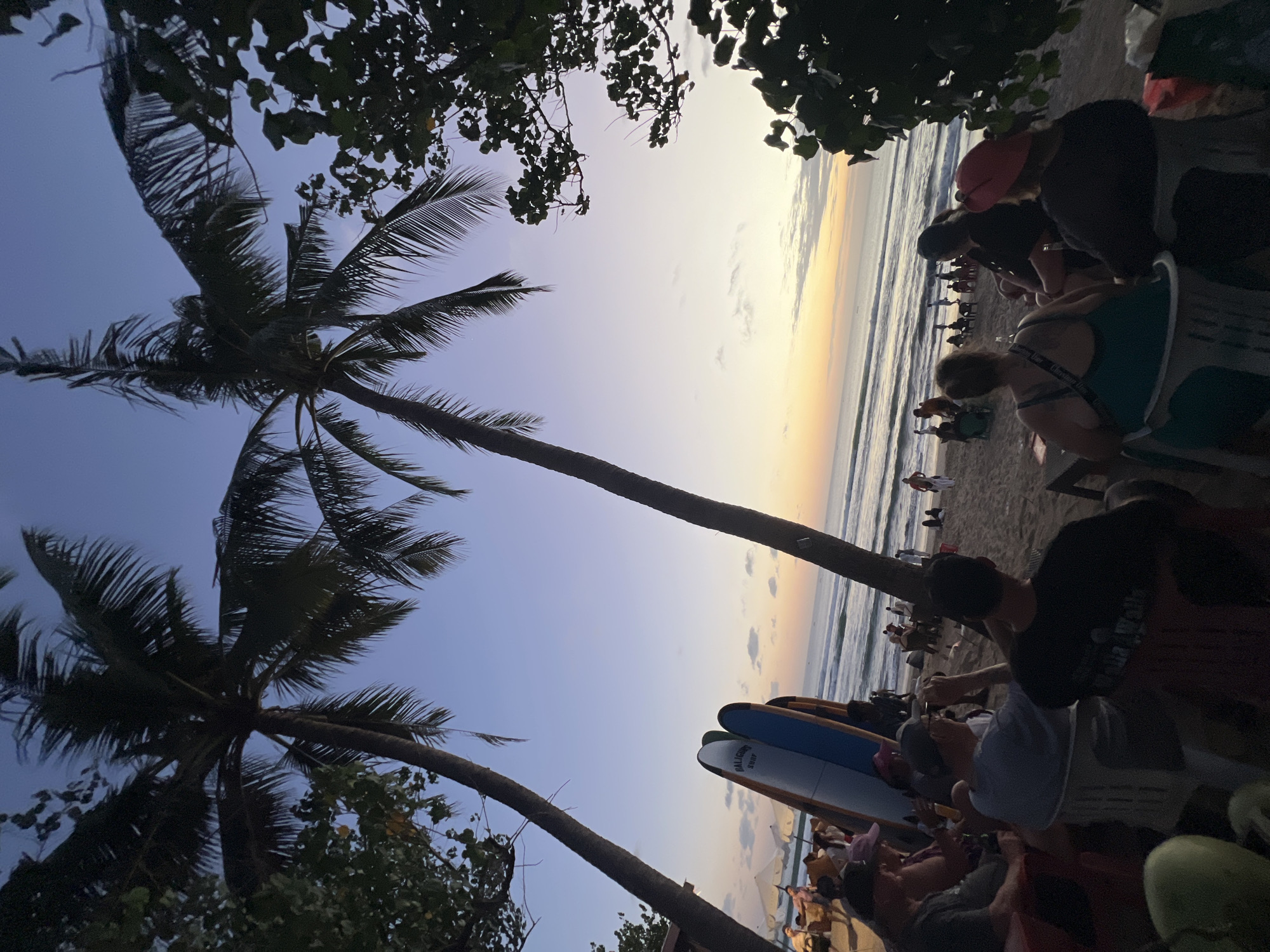 The image depicts a beach scene in Kuta, Indonesia, during twilight. In the foreground, several people are seated in chairs on the sand, facing the ocean. Two surfboards stand upright near the seating area. Palm trees frame the scene, with their fronds silhouetted against the dusky sky. The ocean stretches to the horizon, where a warm glow indicates the setting sun. In the mid-ground and background, more people are scattered on the beach, some walking, others seated at tables. Beach umbrellas are visible in the distance. The overall atmosphere is relaxed and serene, suggesting a popular spot for evening leisure.