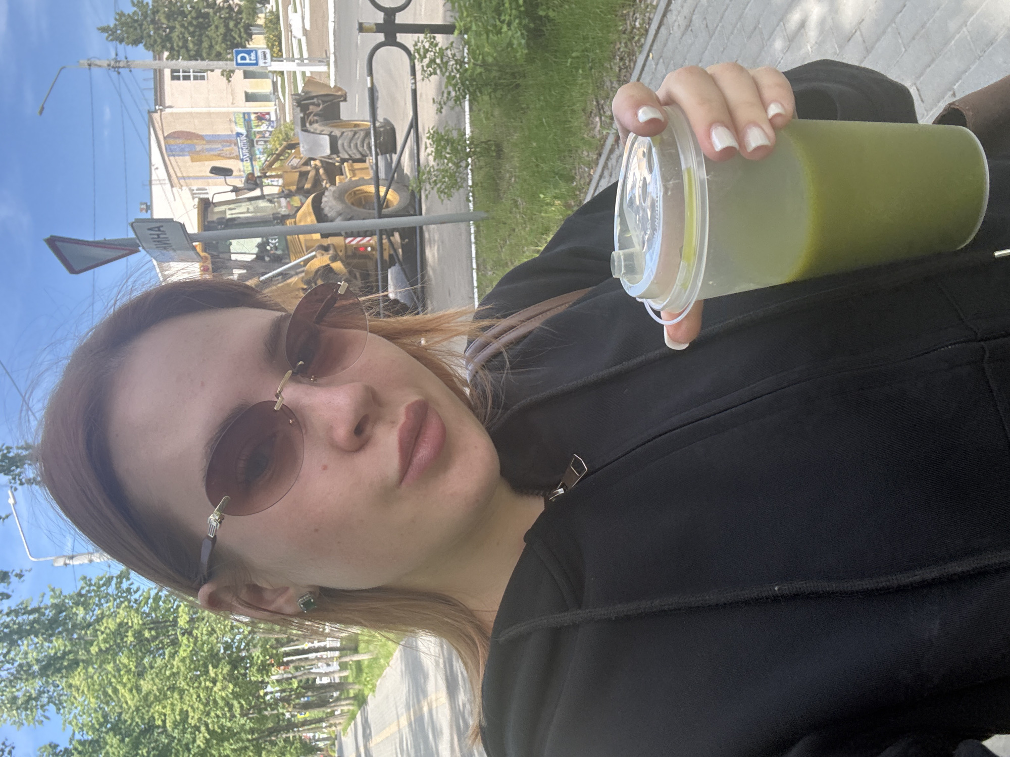 A young woman with reddish-brown hair and sunglasses takes a selfie outdoors on a clear, sunny day. She is wearing a black hooded jacket and has white manicured nails. In her right hand, she holds a clear plastic cup filled with a green beverage, possibly a smoothie or juice. Her expression is neutral and composed.
The background presents an urban or semi-urban street scene. To her left, a bright blue sky is visible, along with overhead power lines and a street lamp. A large yellow construction vehicle, resembling a front-end loader, is parked on the street behind her. Beyond the vehicle, there are light-colored buildings, one featuring a faint mural or sign. Trees and green foliage line the street and pavement. The woman is standing on a paved sidewalk.
Notable details include a white triangular road sign that partially displays the Cyrillic word "ИННА" and a blue parking sign with a white "P". The scene depicts a casual daytime moment in what is identified as Chaikovsky, Russia.