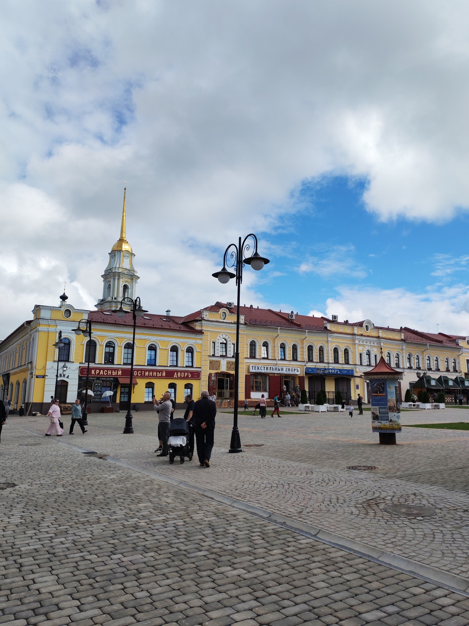 The image shows a wide cobblestone plaza in front of a yellow, multi-story building with a red roof. The building features arched windows and several signs, including "КРАСНЫЙ ГОСТИНЫЙ ДВОР" (Red Merchant Courtyard) and "ТЕКСТИЛЬНЫЙ ДВОР" (Textile Courtyard). A tall, slender golden spire with a cross atop it rises from the center of the building. Several streetlights with decorative curly arms are positioned throughout the plaza. A few people are visible walking, including one person pushing a stroller, and another taking a photo. The sky is partly cloudy with patches of blue. The scene appears to be in the city of Bol'shie Mkhi, Russia.